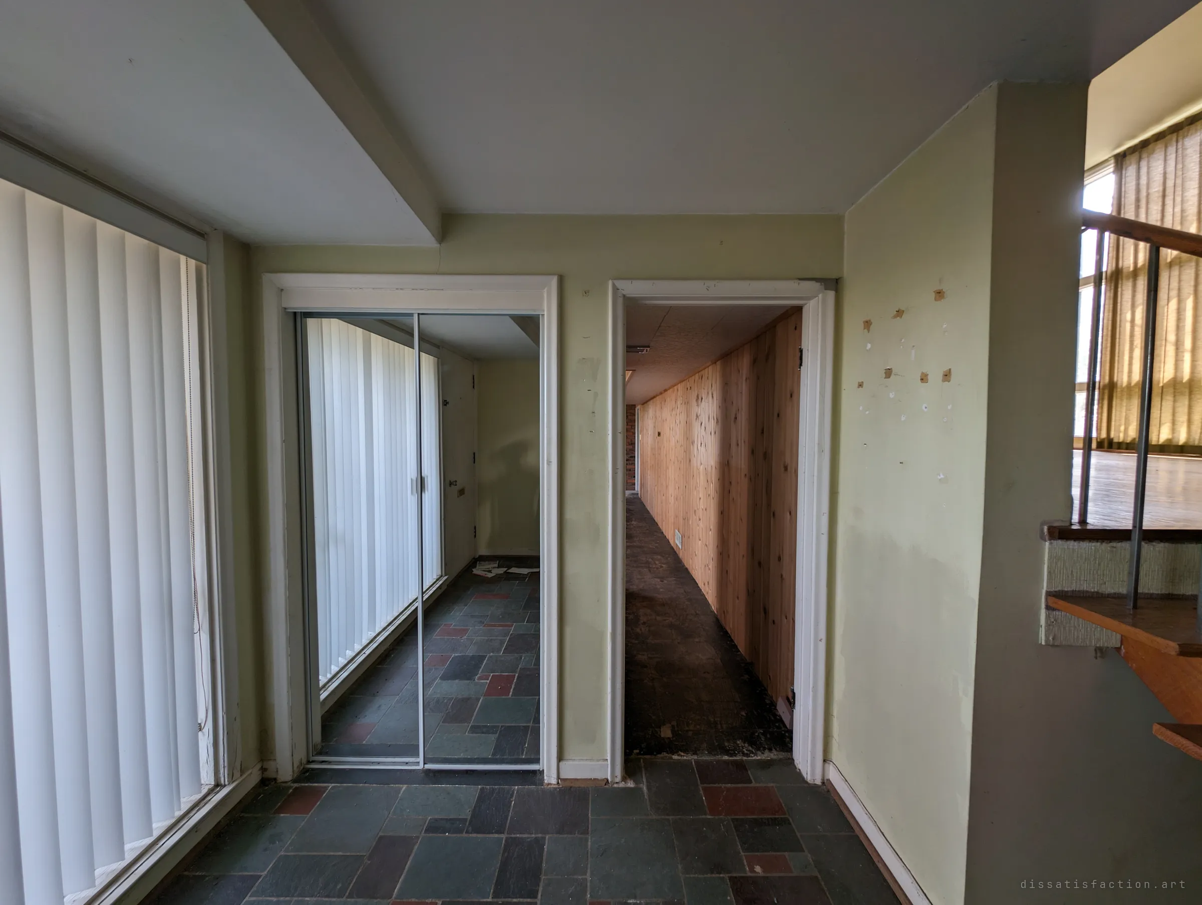 A slate-tiled landing with vertical blinds, a mirrored closet door and a cedar-panelled corridor stretching away through the next doorway. A glimpse of a sunlit open room past the stair rail on the right shows the house's split levels.