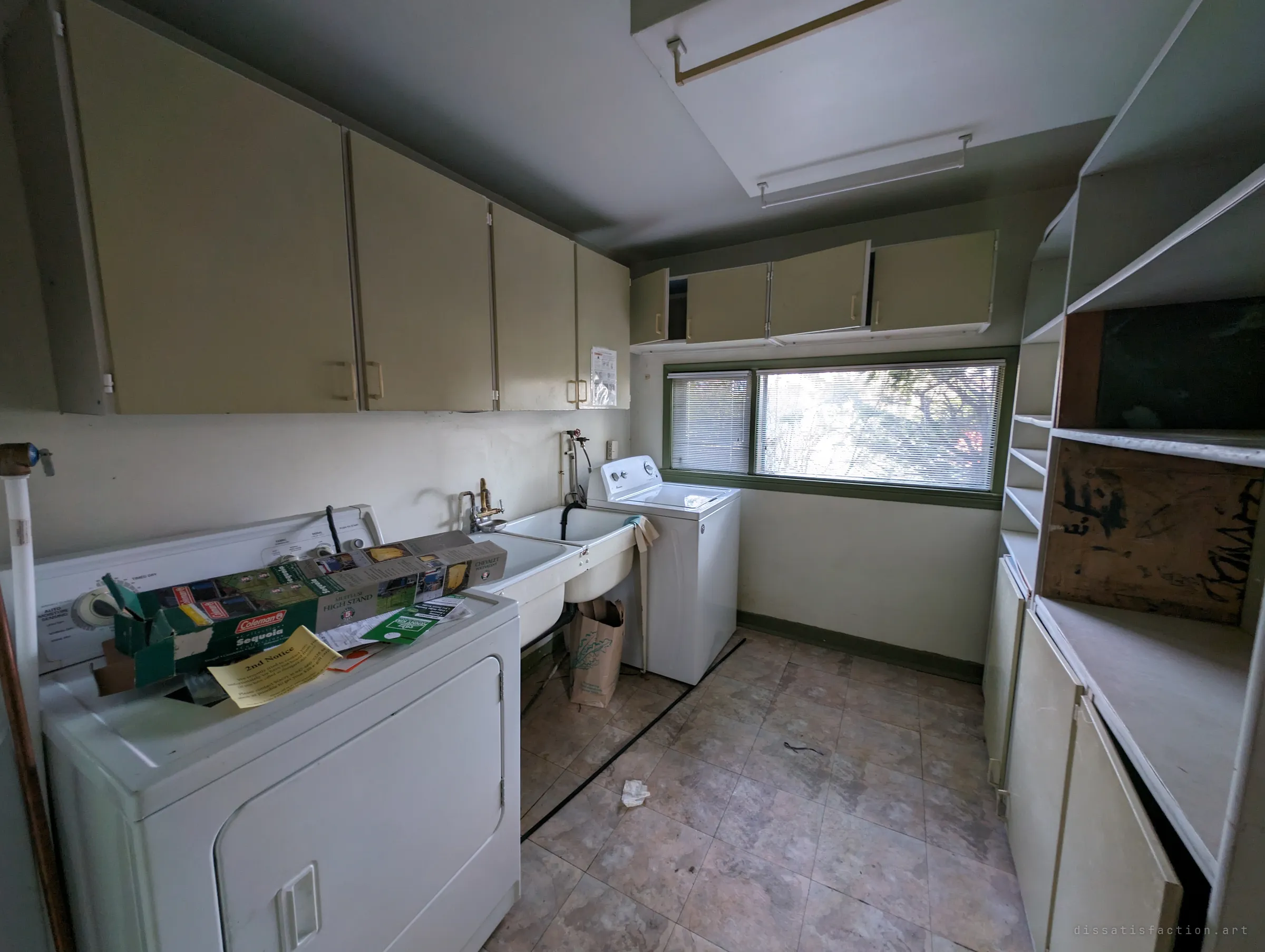 A laundry room with a dryer, twin utility sinks and a washer under a blind-covered window, cabinets lining the walls. Leftover mail and a boxed camping stand on the dryer suggest the owners left in a hurry.