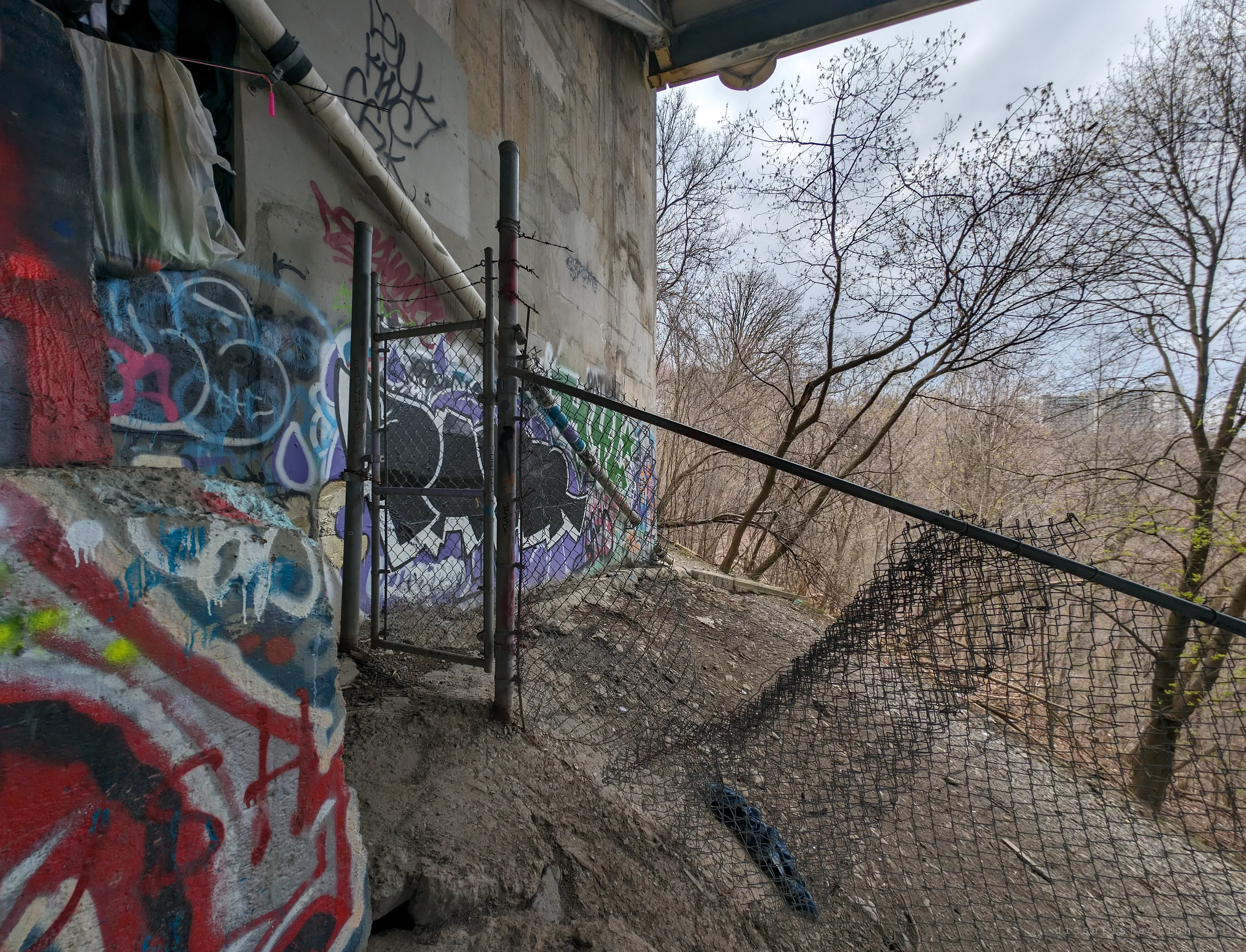 A graffiti-covered bridge abutment with a barbed-wire gate and a torn chain-link fence hanging over a leafless ravine. Someone has strung plastic sheeting inside the ledge, a sign the spot is lived in.