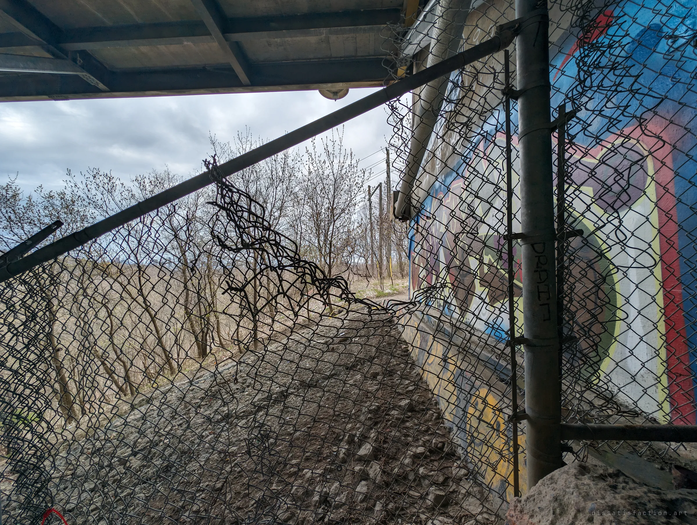 Looking through a ripped chain-link fence under the deck edge, a narrow gravel ledge runs past a painted wall toward bare trees and utility poles. The mangled mesh shows how often the barrier has been forced.
