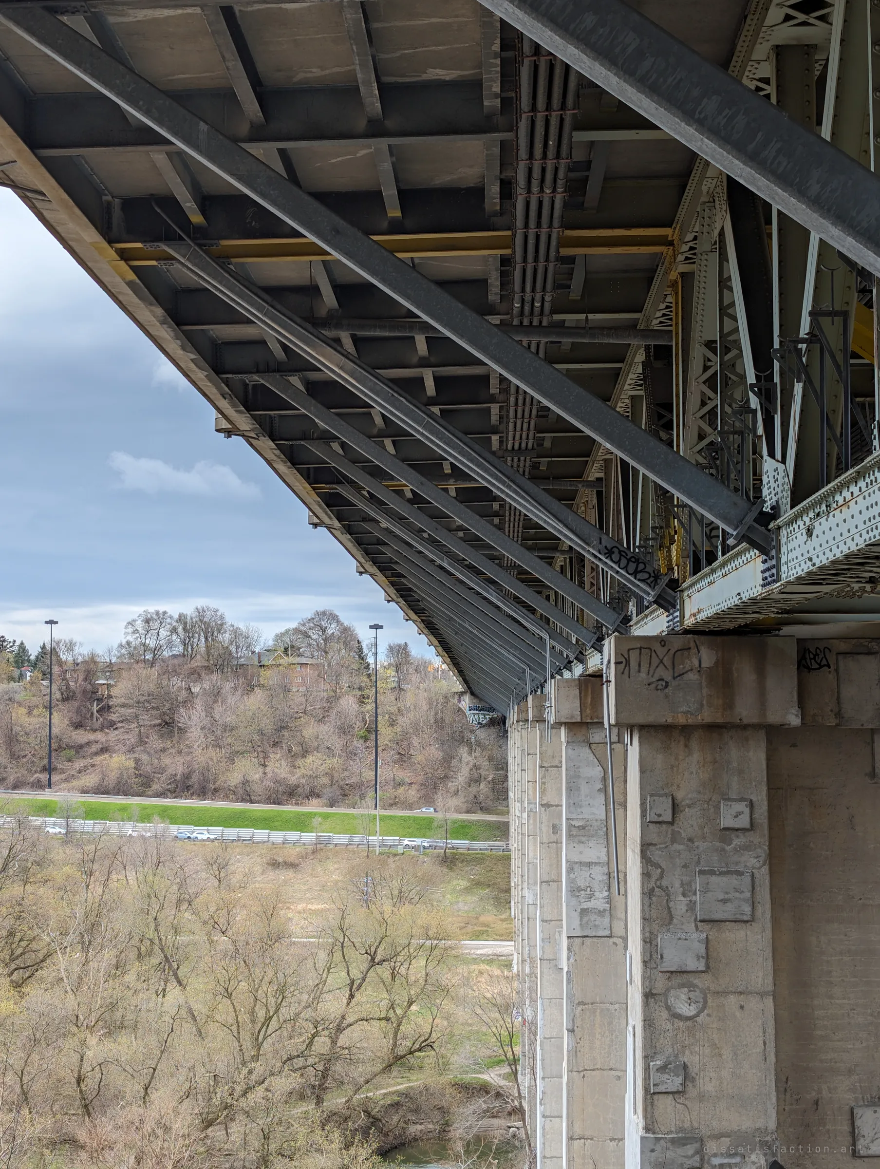 The underside of a long steel deck stretches away on a row of concrete piers above a valley of bare trees and a highway. It shows the sheer scale of the structure from beneath.