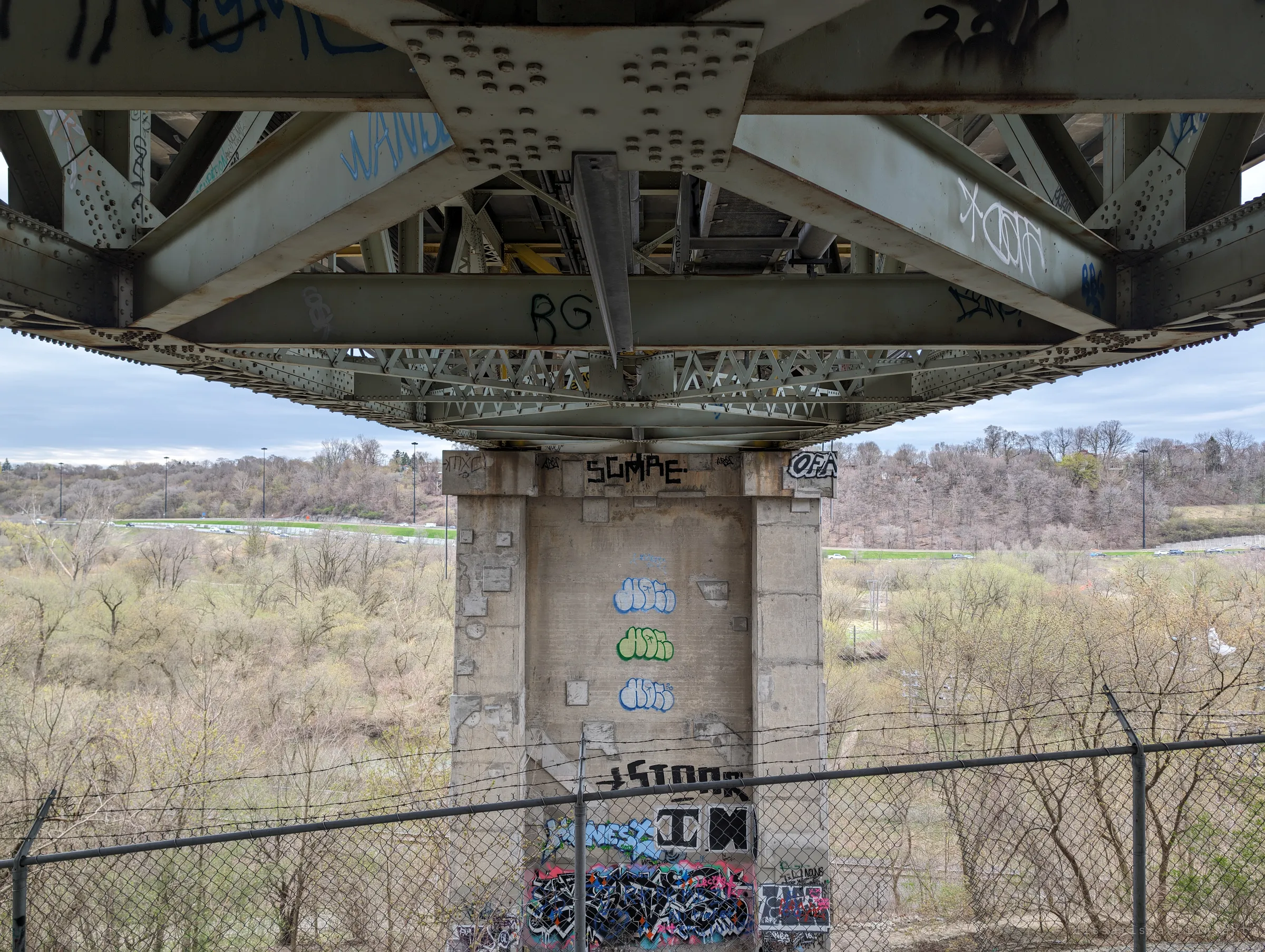 A riveted steel truss sits on a tagged concrete pier, with a barbed-wire fence in the foreground and a wide valley behind. The symmetry of the steelwork frames the graffiti-stacked column.