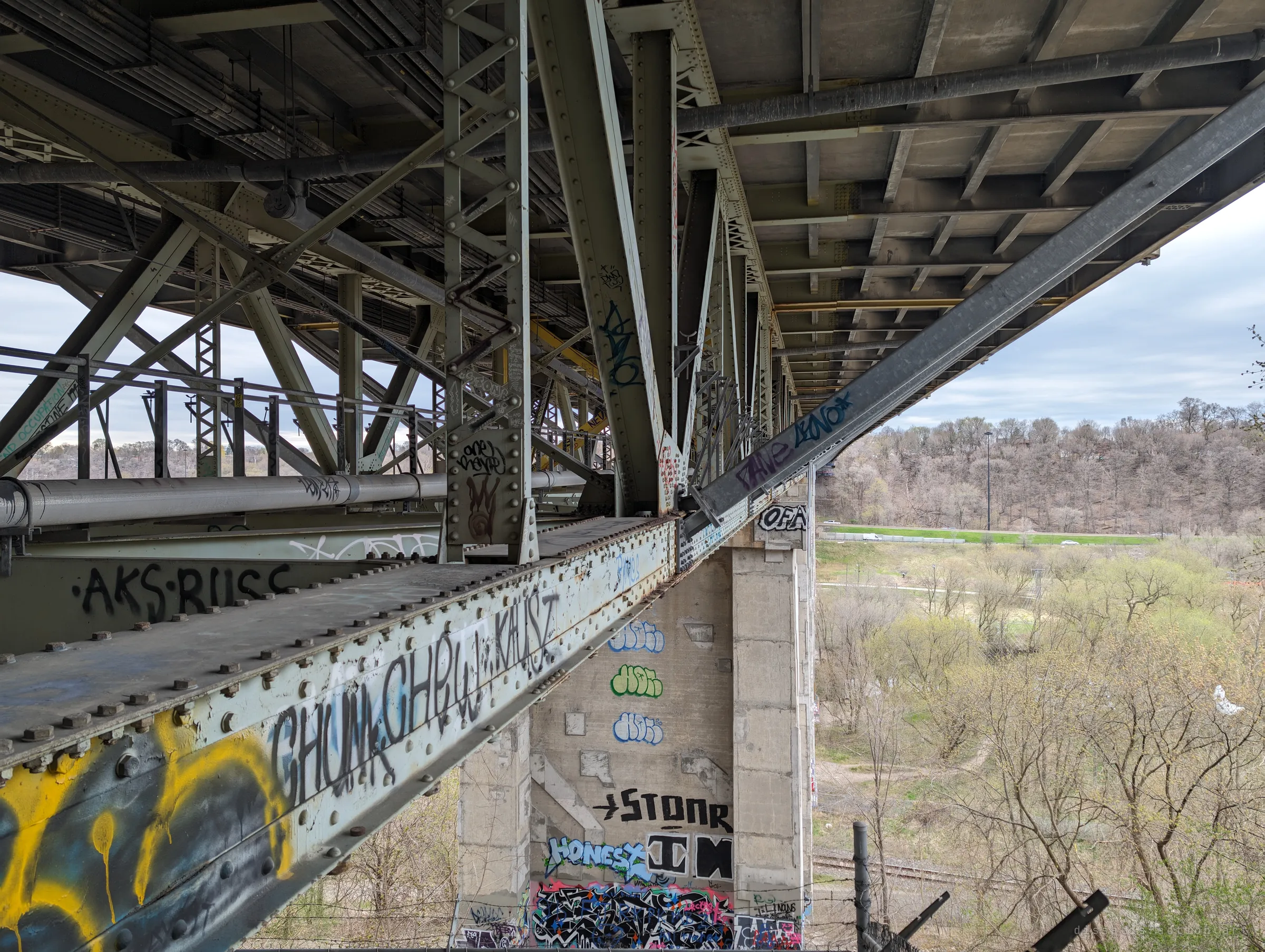 A graffiti-coated steel girder runs diagonally into a web of trusses and a concrete pier, with the valley opening up on the right. The tags reach even the high, hard-to-get-to beams.