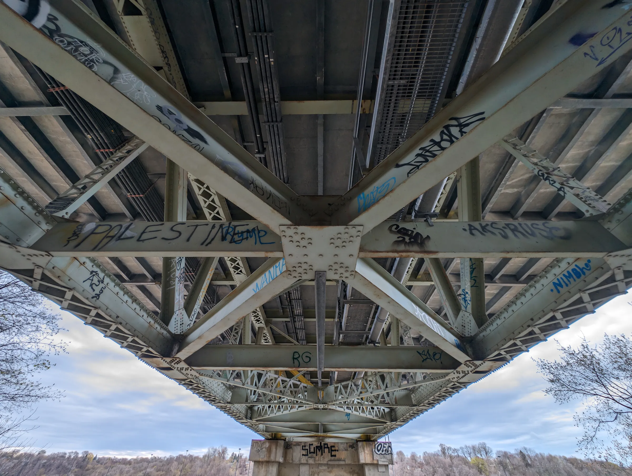 Looking straight up at the underside of the span, tagged steel cross-bracing converges on a riveted gusset plate above a single pier. The view turns the structure into a symmetrical pattern.
