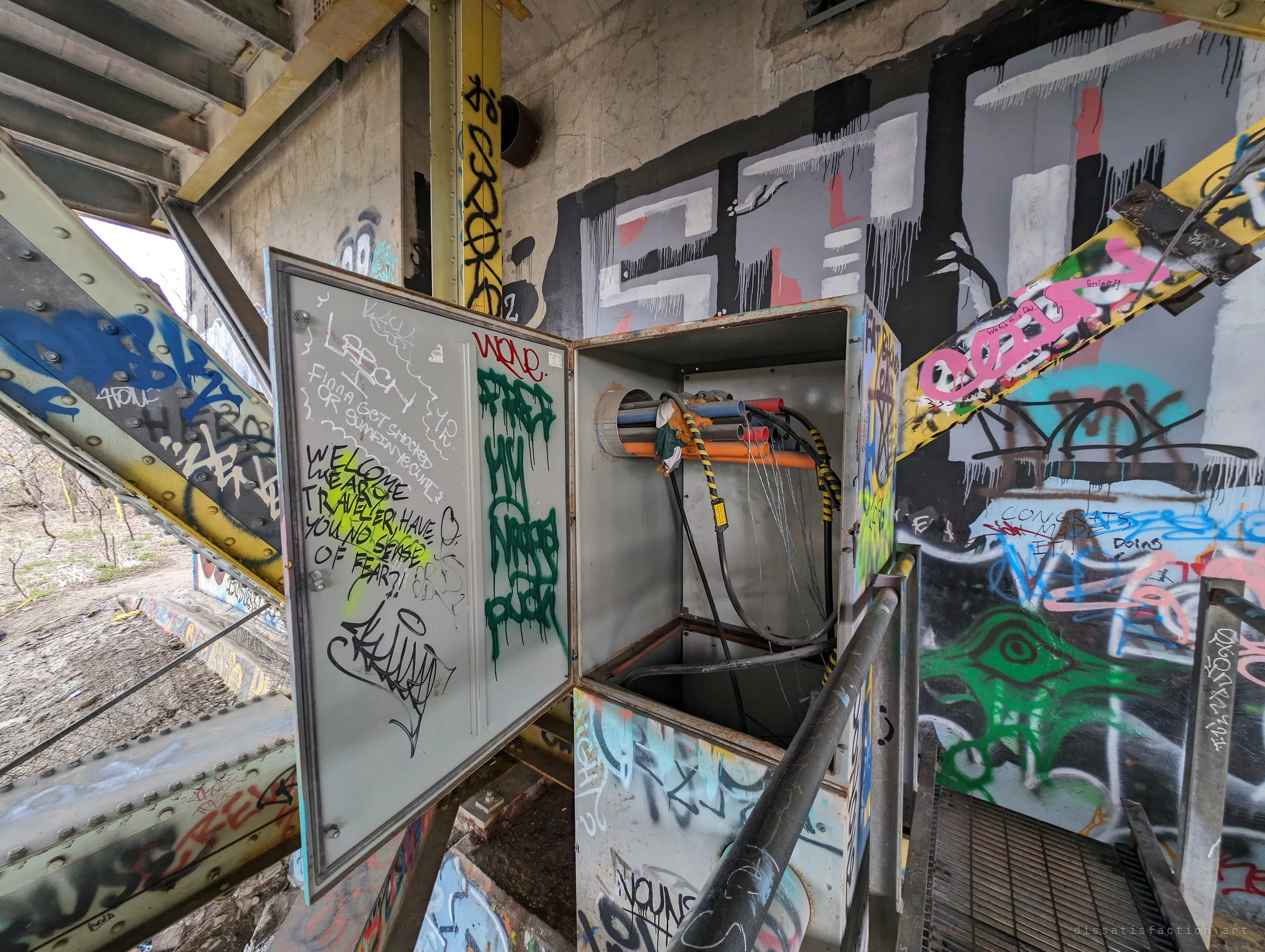 An open electrical cabinet, its wiring gutted, stands on a catwalk inside the bridge's abutment chamber surrounded by graffiti. A scrawled message on the door warns visitors about their lack of fear.