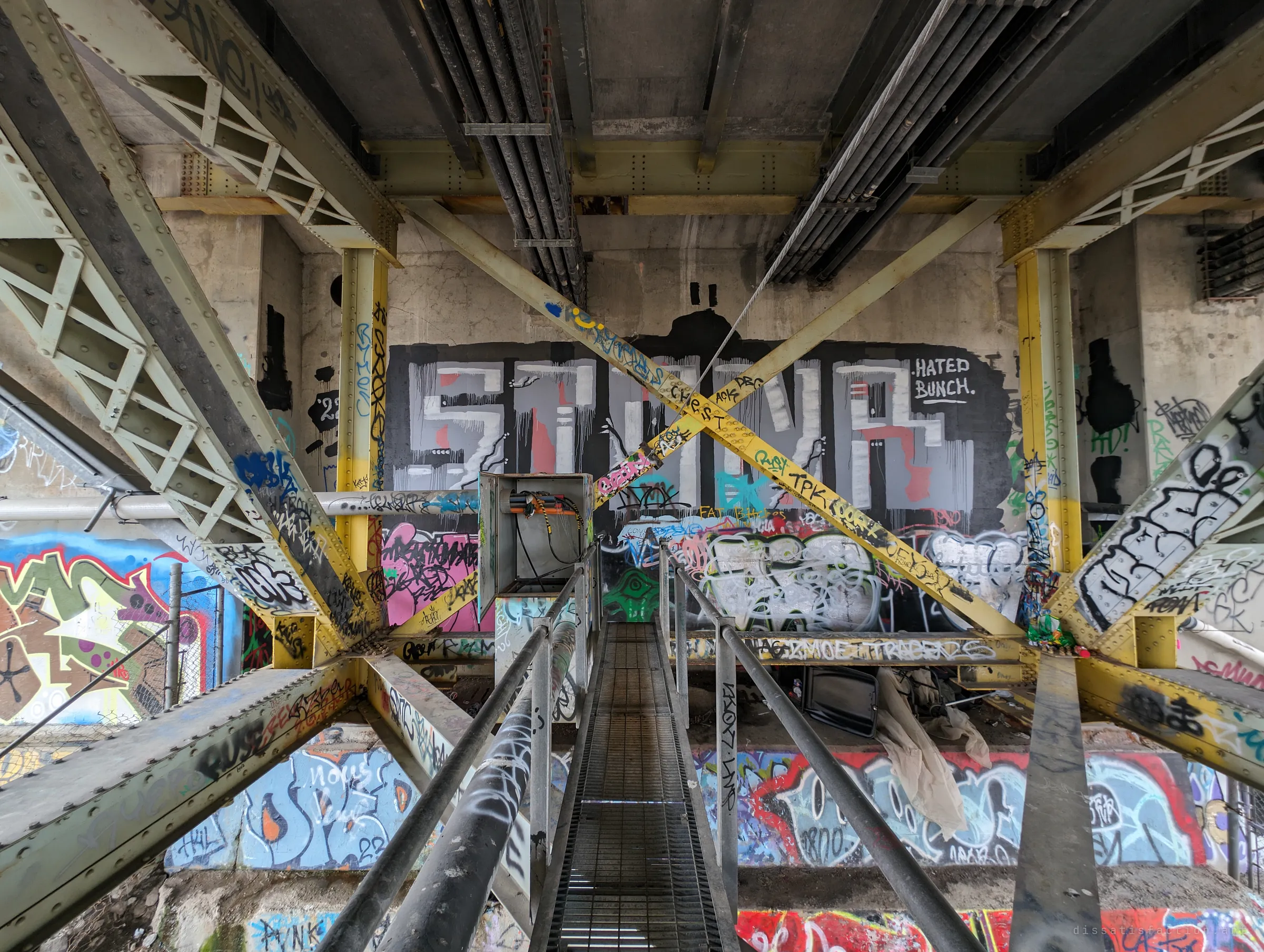 A grated catwalk leads between yellow cross-braces toward a wall-sized graffiti piece inside the abutment. Discarded clothes and debris below show people shelter here.