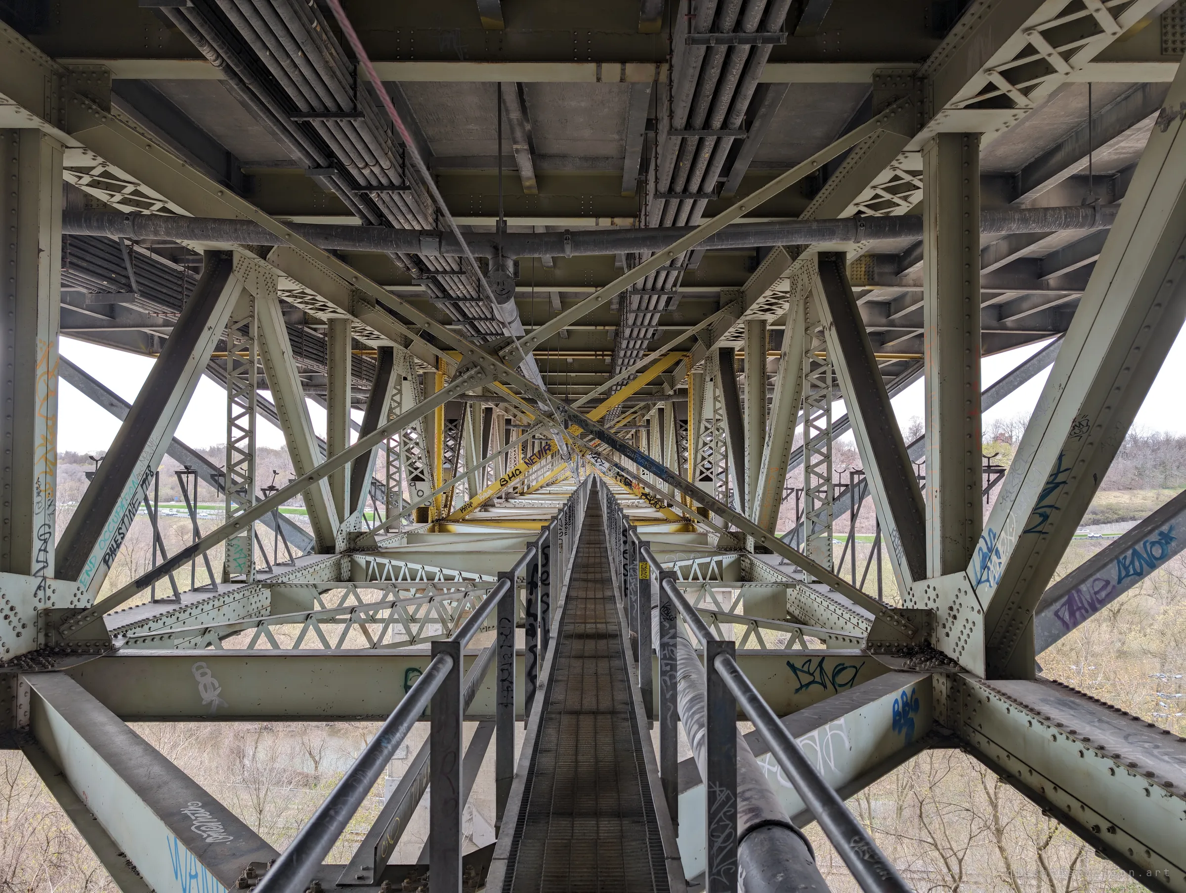 A narrow inspection catwalk runs dead centre through the bridge's steel trusses, vanishing toward a far vanishing point. The repeating bracing makes the length of the span tangible.