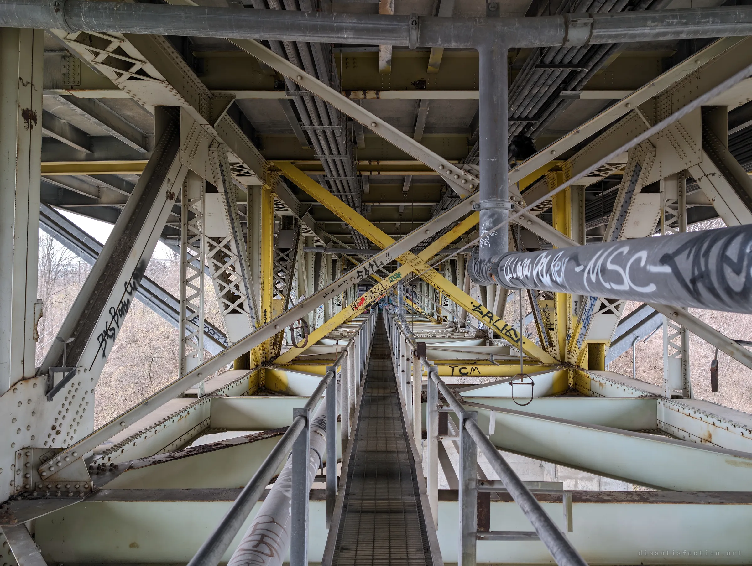 From deeper along the catwalk, yellow X-bracing and a tagged pipe frame the grated walkway stretching ahead under the deck. The enclosed tunnel of steel feels far removed from the valley outside.