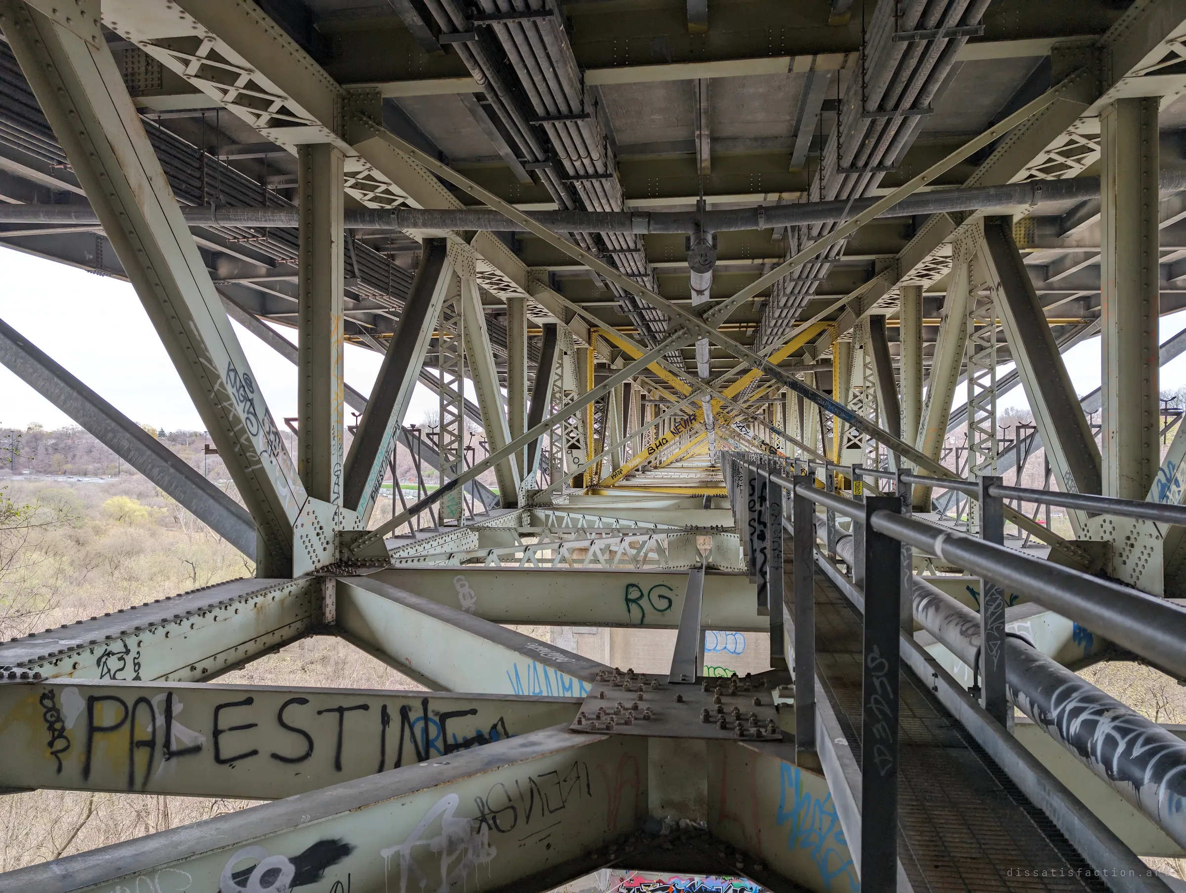 Looking down the length of a steel truss bridge's underside, a narrow catwalk with railings running into a tunnel of riveted girders and graffiti. The repeating X-bracing pulls the eye toward a vanishing point far above a leafless ravine.
