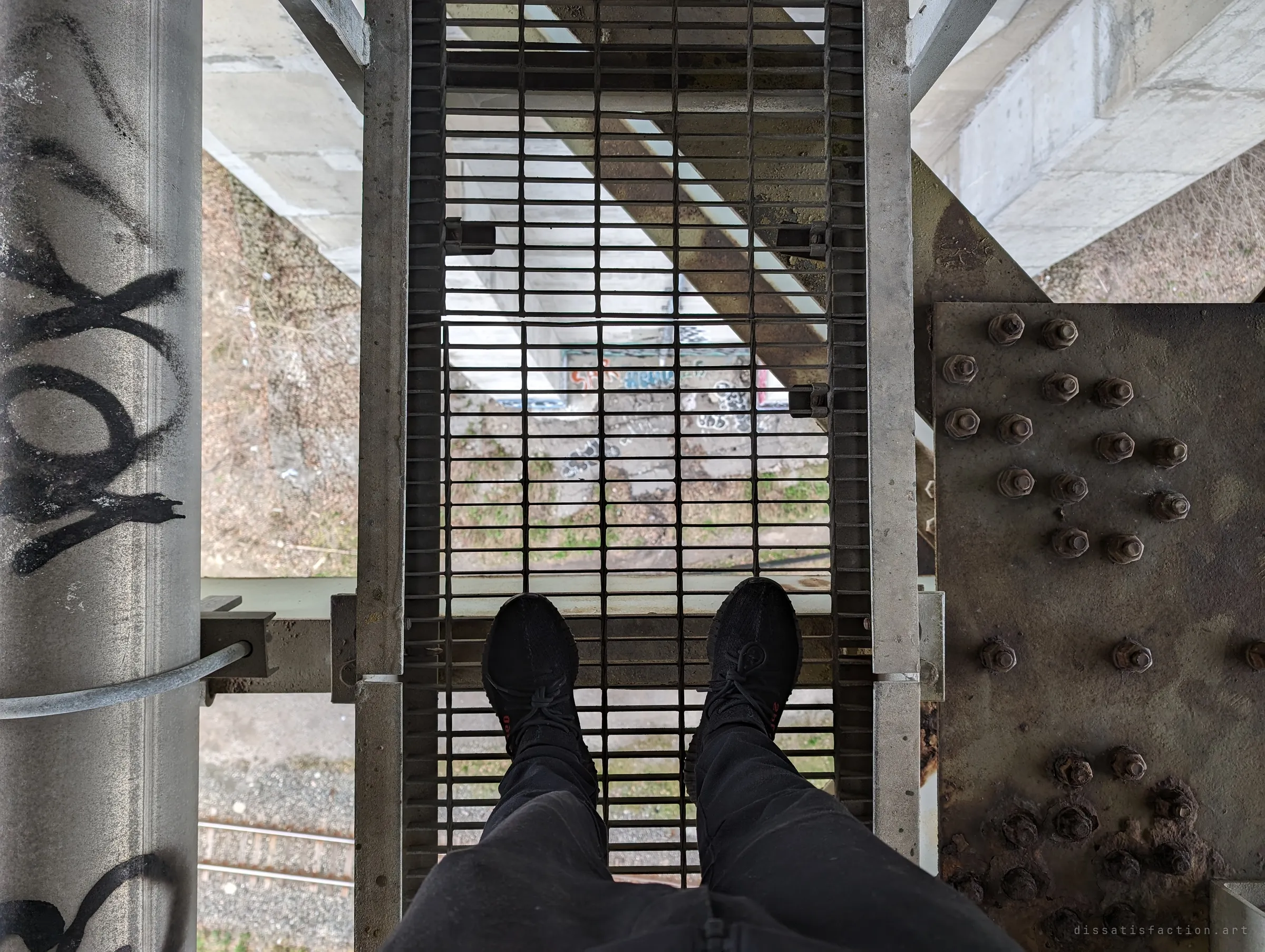 Feet in black sneakers standing on open steel grating, with the ground, rail tracks and a graffitied pier far below. It shows just how exposed the catwalk is.