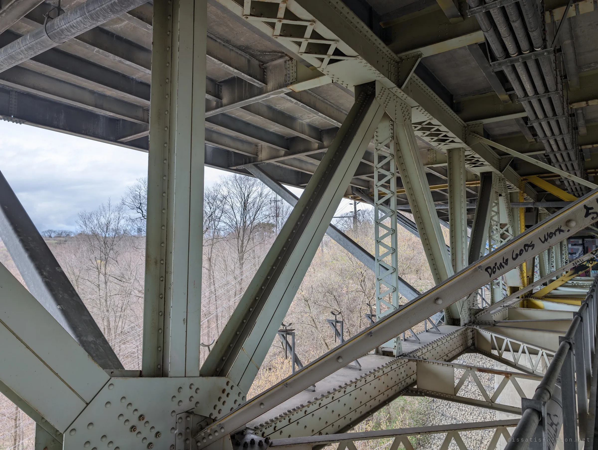 Pale green riveted steel beams and lattice columns fan out beneath the deck, framing bare spring trees on the ravine slope. Hand-written graffiti runs along one diagonal brace.