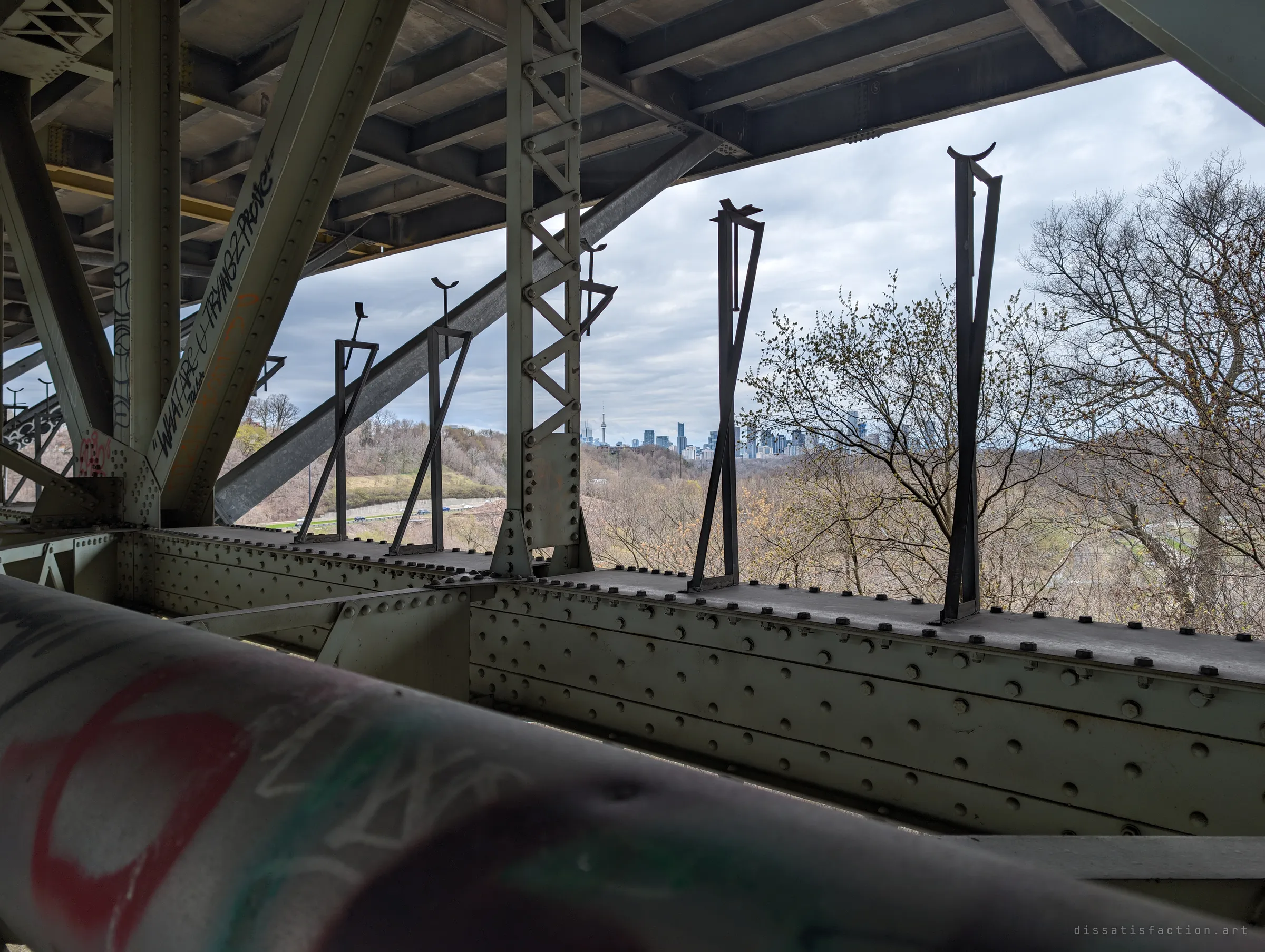 Standing inside the truss, a graffitied pipe blurs across the foreground while riveted girders and tall steel brackets frame a distant downtown skyline beyond the treetops. The city feels far away from up here.