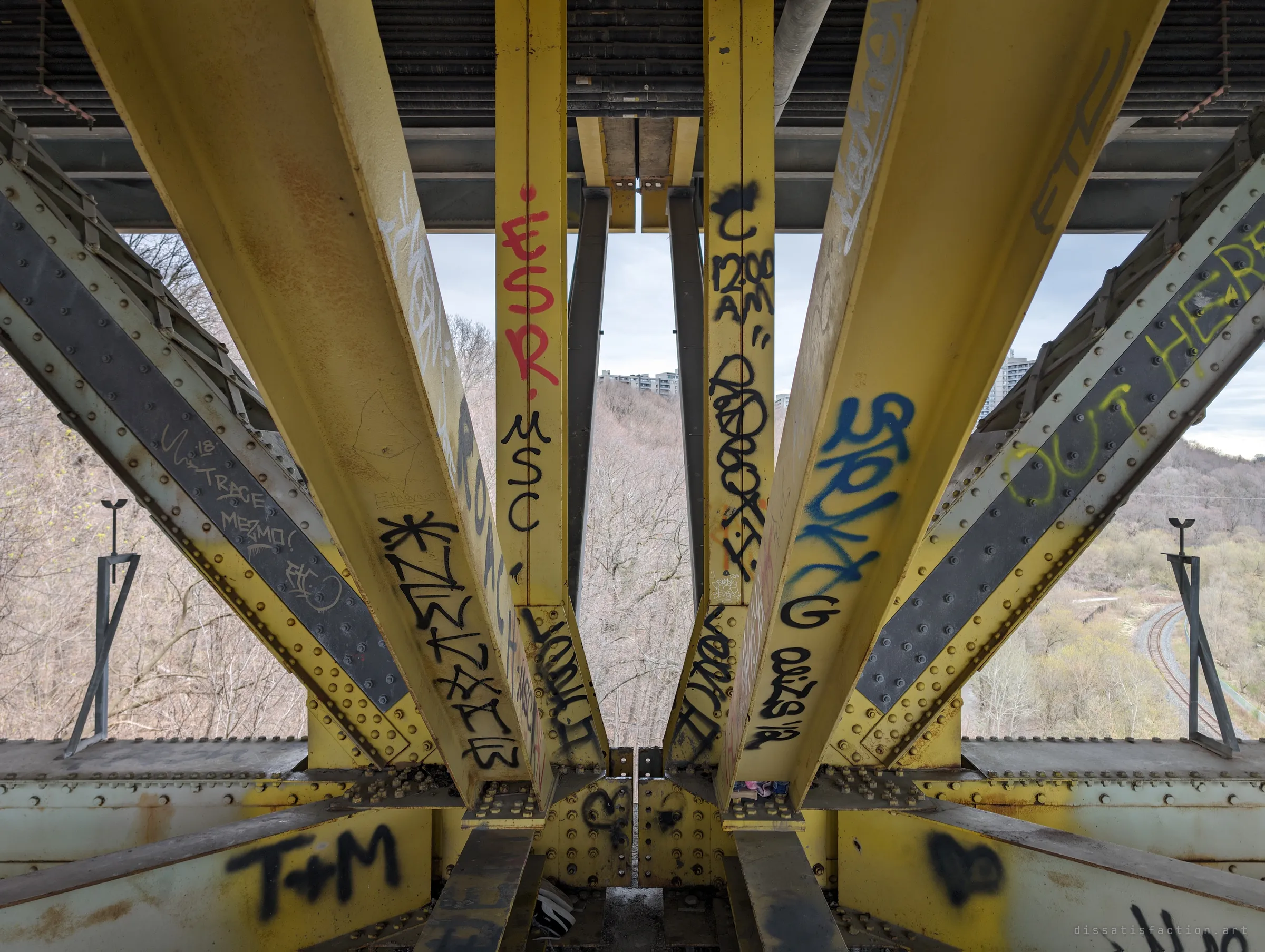 A symmetrical view between yellow steel columns and diagonal braces thick with graffiti tags, opening onto the wooded ravine below. The bridge's skeleton converges like a church nave.