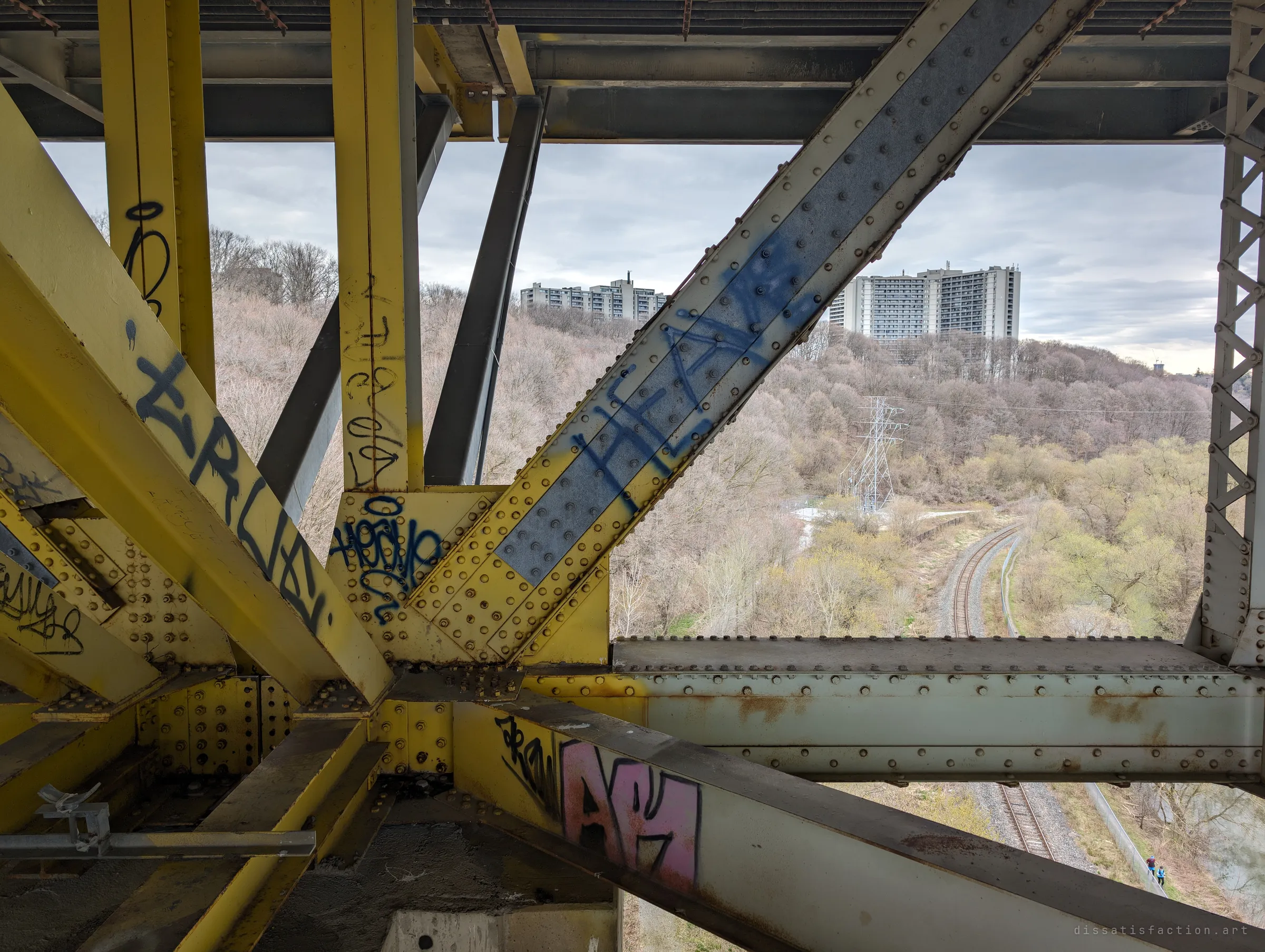 Yellow and pale green riveted girders cross in the foreground, tagged in blue and pink, while the ravine opens out beyond with rail tracks curving through the trees. Two tiny figures walk the path far below.