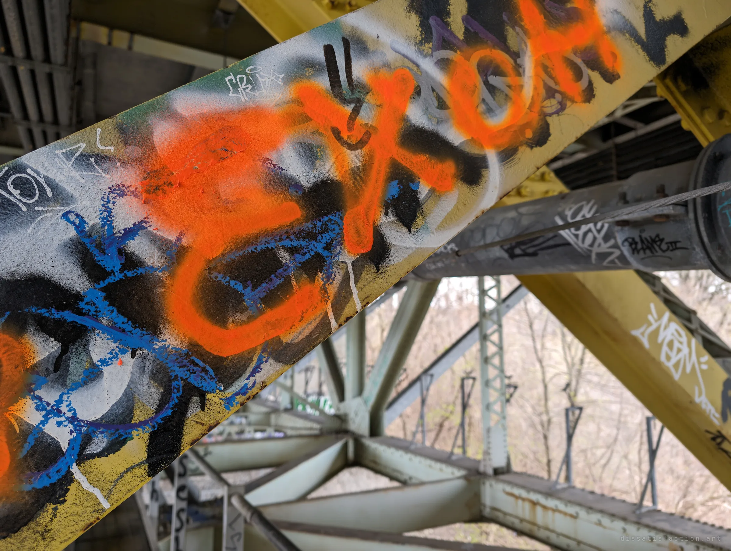 A close-up of a yellow brace layered with orange, blue, black and white graffiti, the truss and bare trees soft behind it. Years of tags stacked on top of each other.