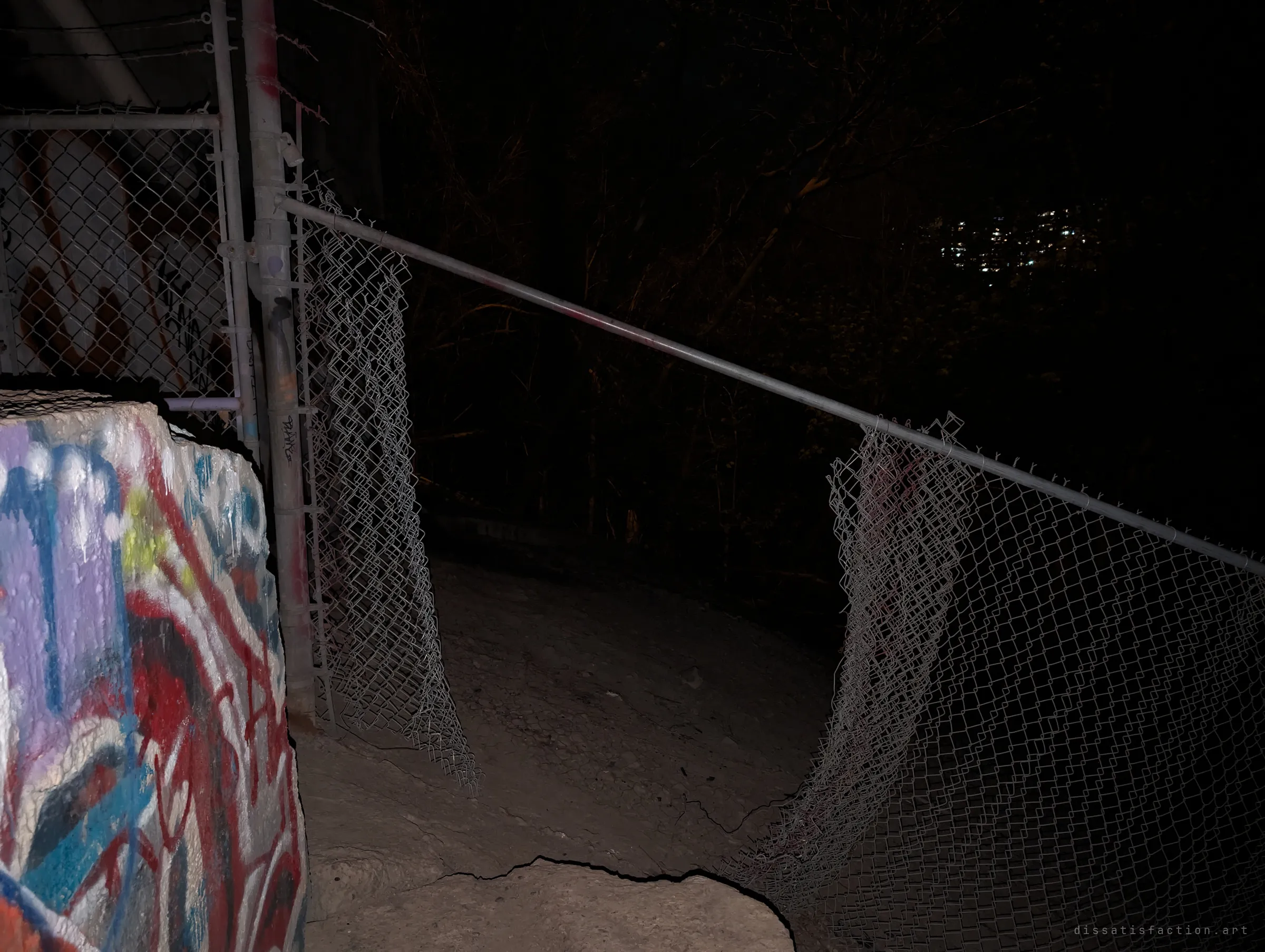A cut and peeled-back chain-link fence beside a graffiti-covered concrete block opens onto a dark wooded slope at night, the way in.