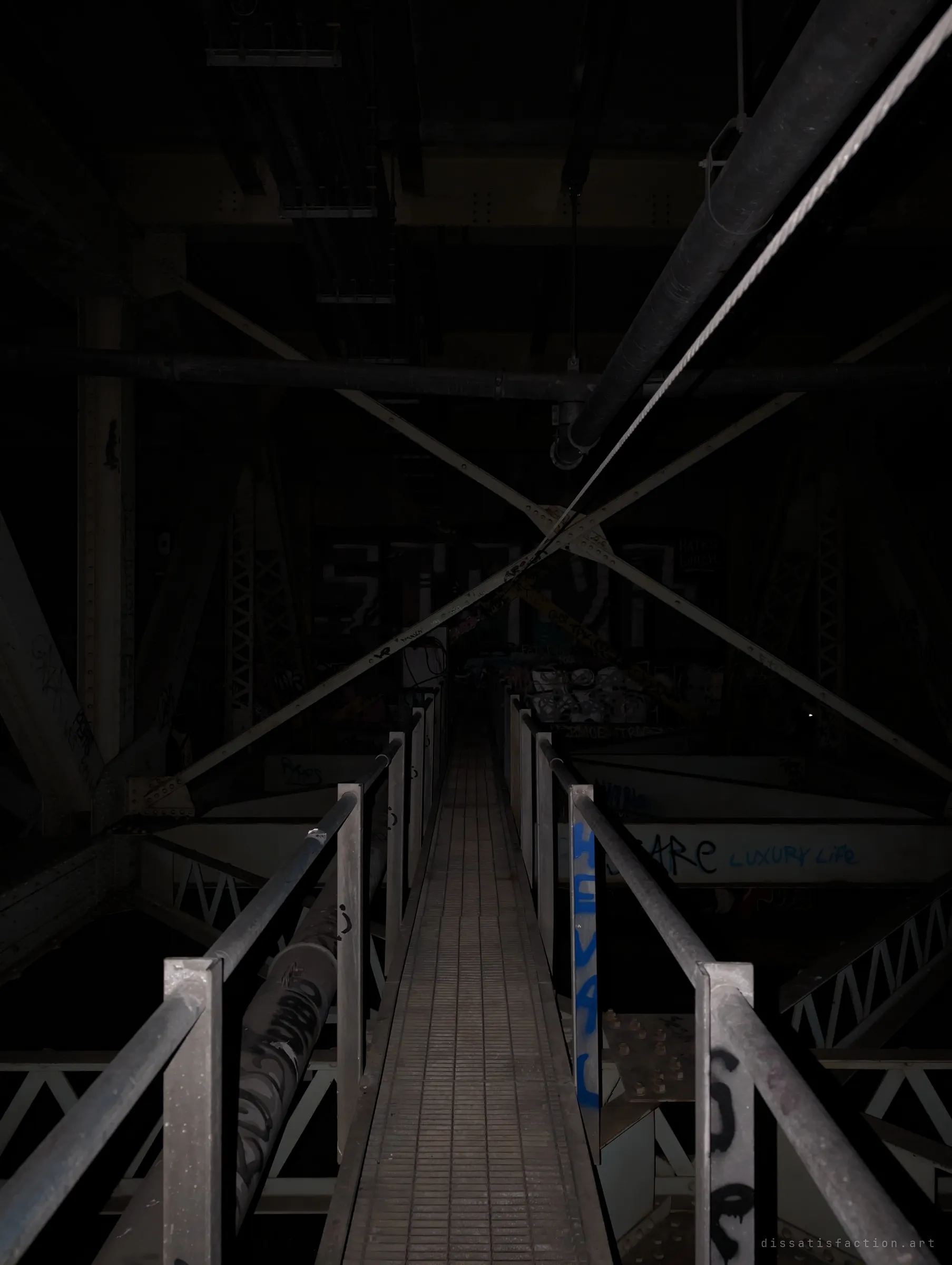 A narrow grated catwalk runs straight into the dark beneath the bridge deck, flanked by graffiti-tagged rails and crossed steel bracing.
