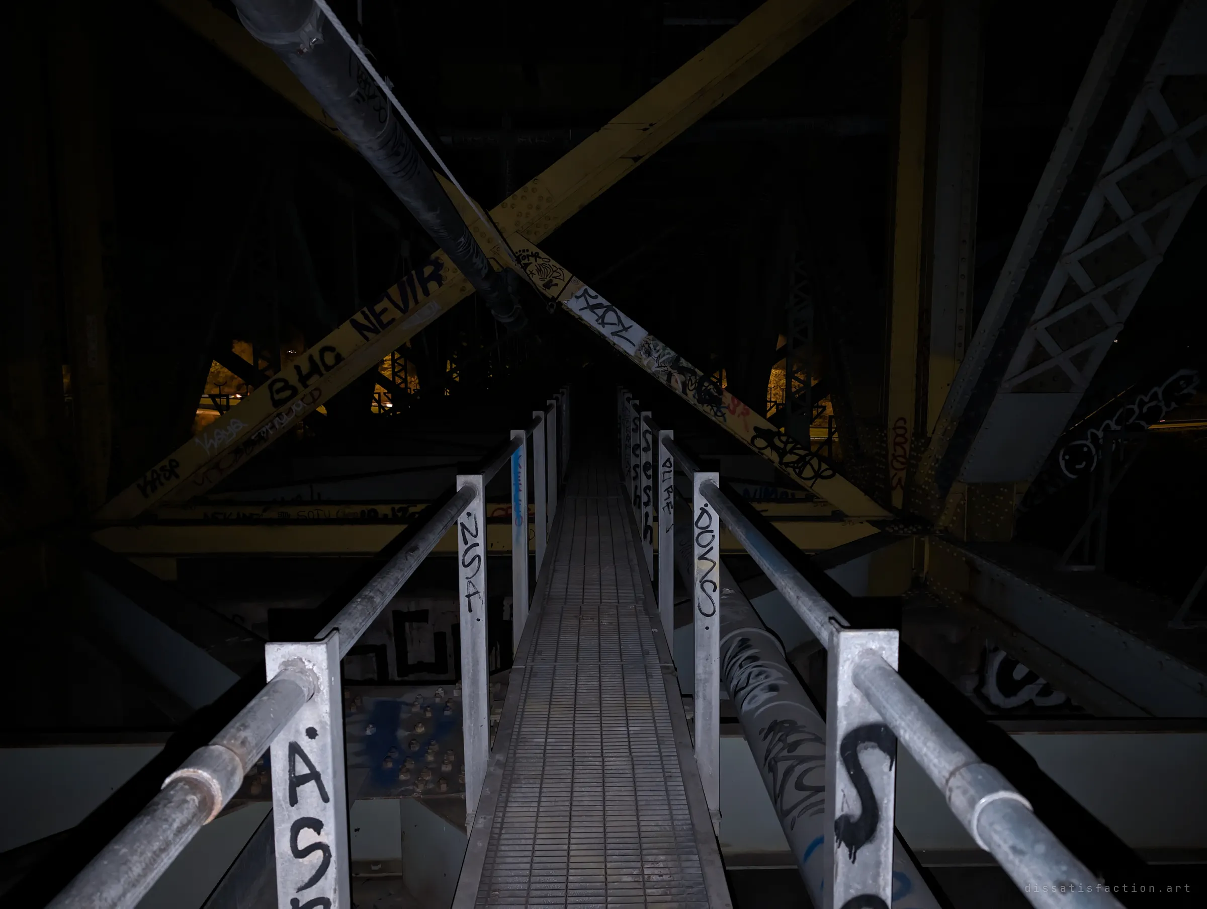 The same catwalk lit by flash, its posts and pipes thick with tags under a giant yellow X of riveted cross-bracing.