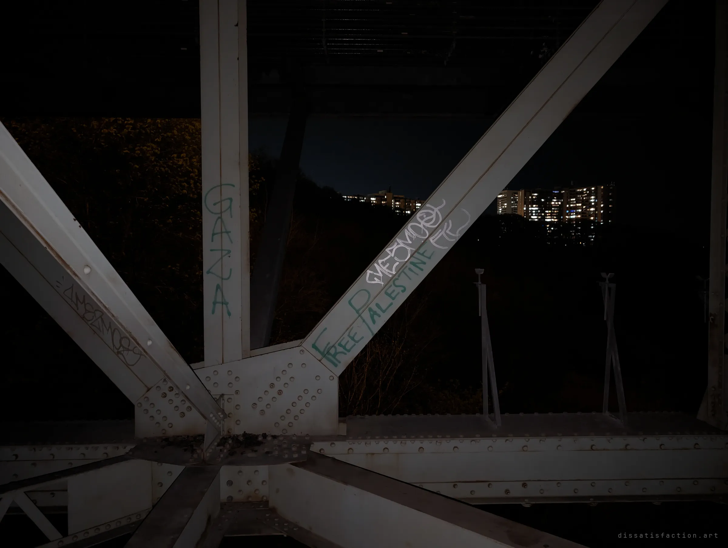 Riveted steel truss members painted with political graffiti frame a night view over the dark ravine to lit apartment towers.