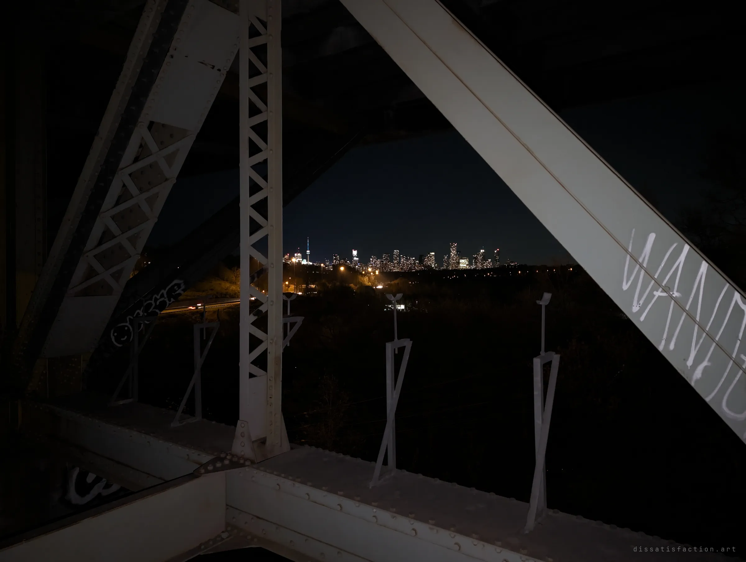 From inside the steel truss, the lit downtown skyline glows on the horizon past a dark valley and a thin line of highway traffic.