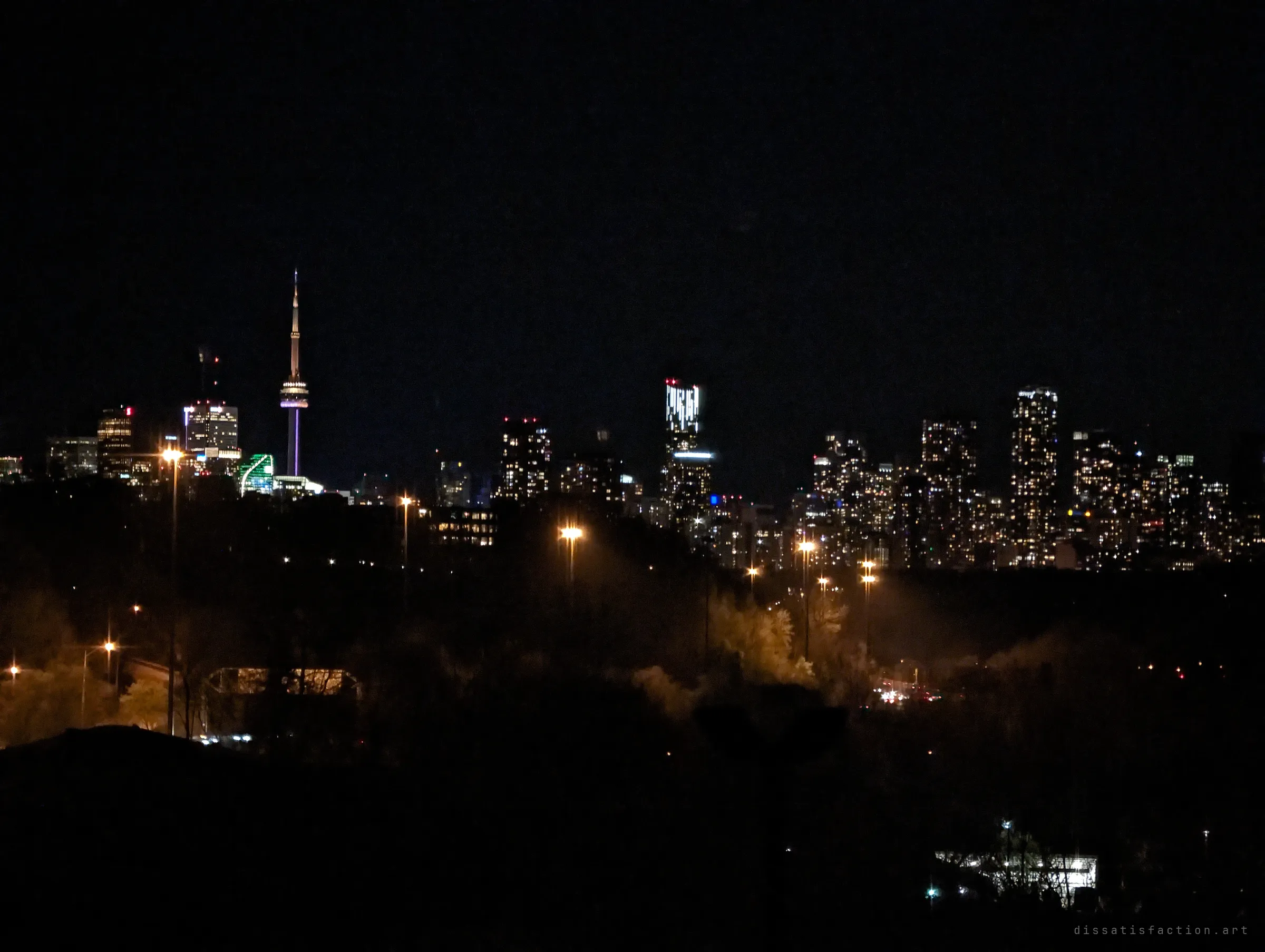A zoomed night skyline with a slender tower and lit condo blocks above sodium streetlights in the valley below.