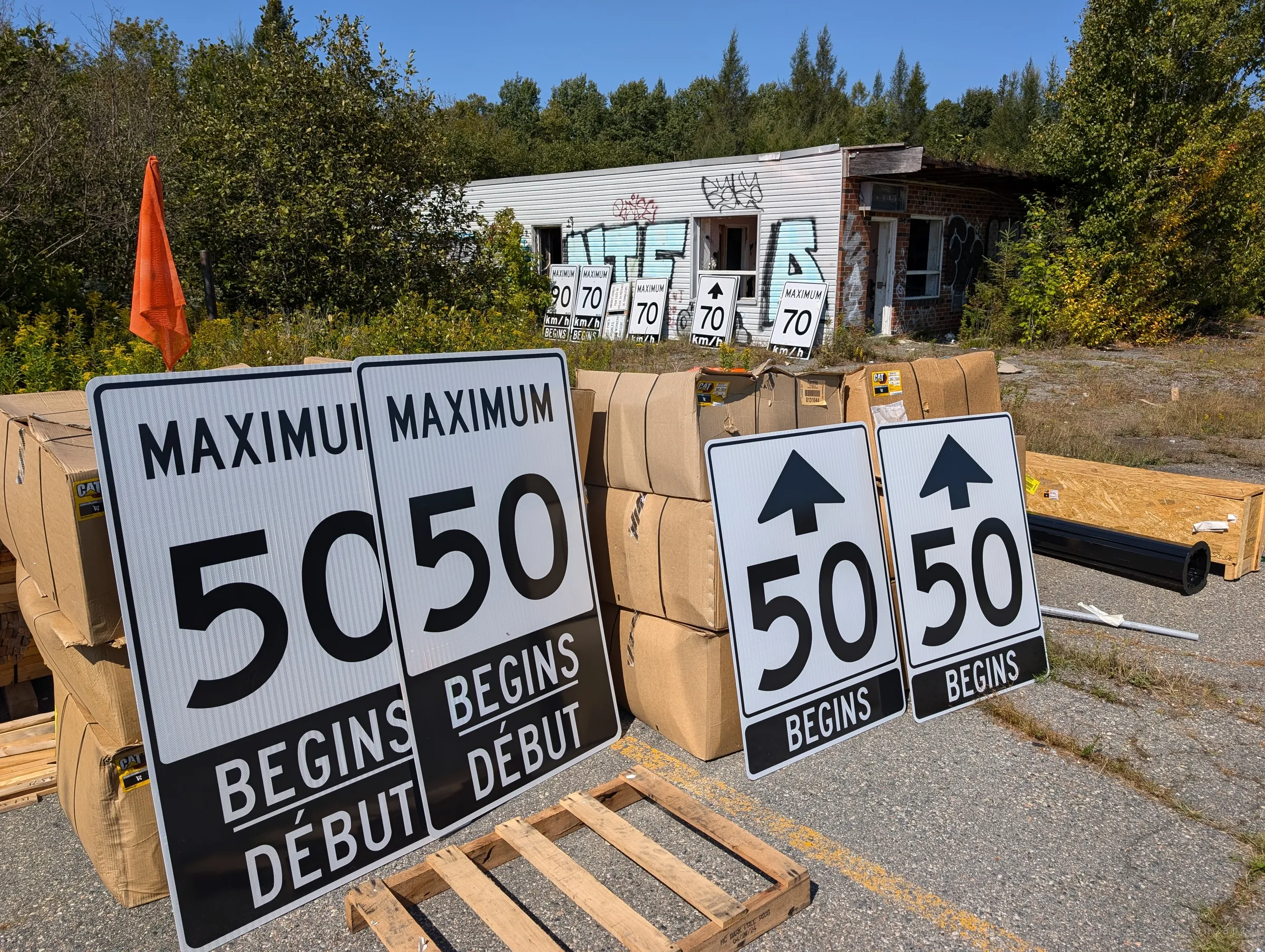 Stacks of brand-new speed limit signs lean against boxed supplies in the cracked lot, with more signs lined up against the graffiti-covered building behind. The abandoned lot has become someone's storage yard.
