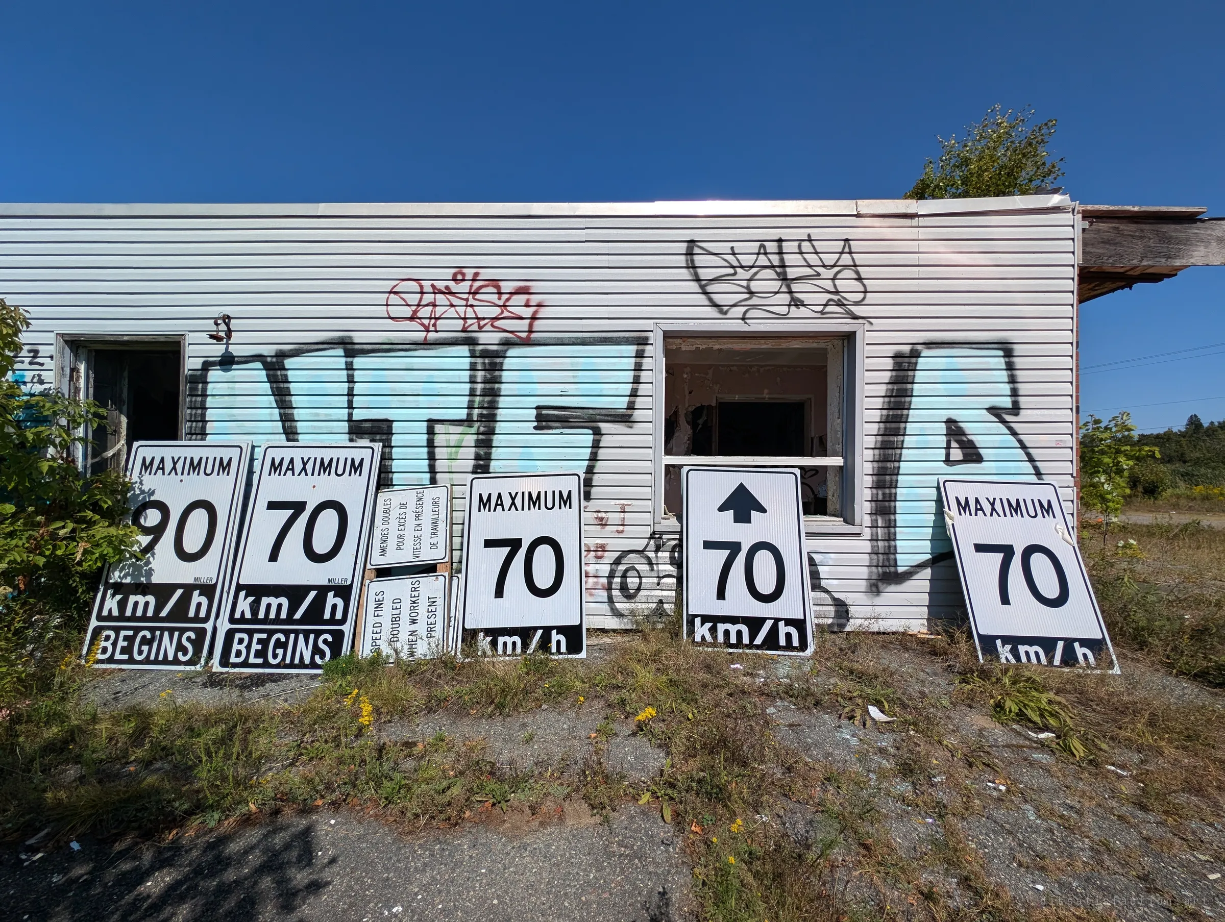 A row of speed limit and work-zone signs leans against a sided wall covered in large graffiti pieces, below a window with the glass gone. Road-crew gear stashed at a dead building.