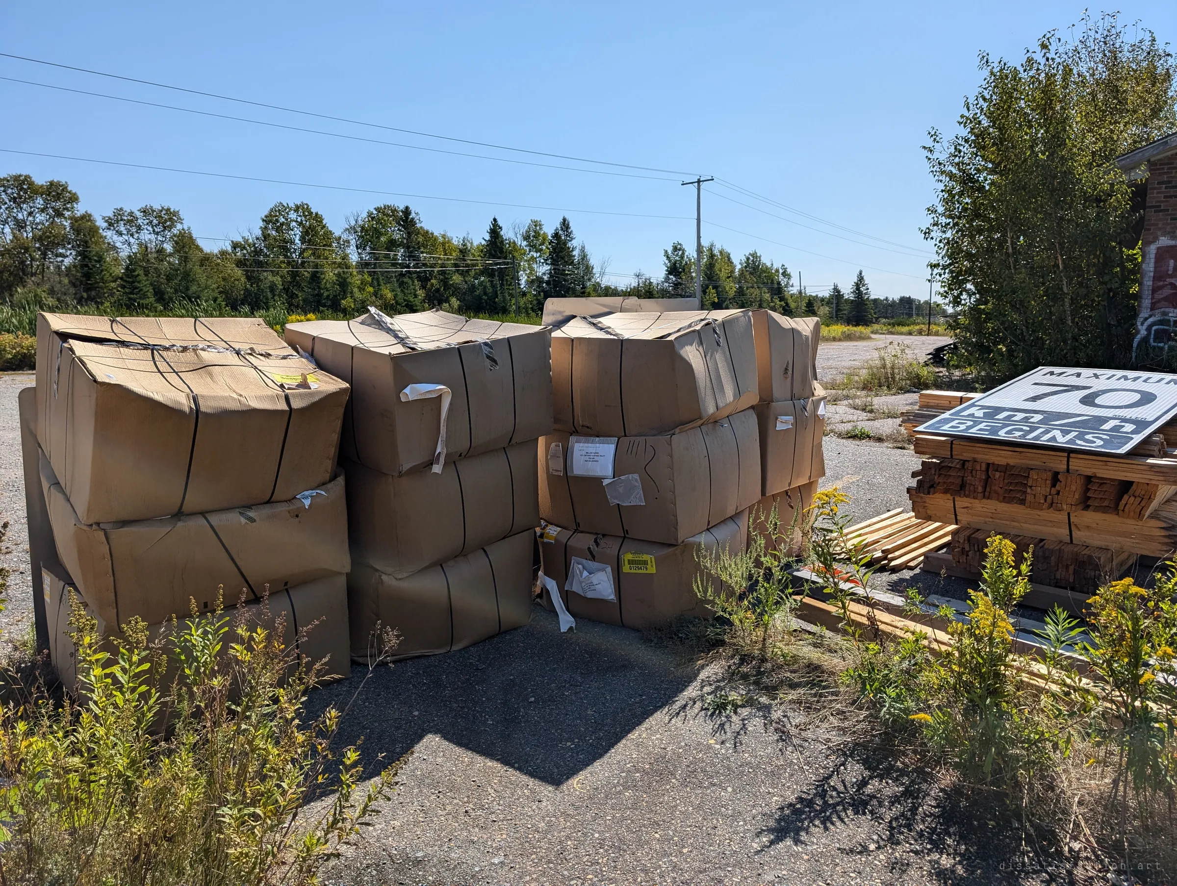 Strapped cardboard crates sit stacked on the old asphalt beside a pile of lumber and a spare speed sign, weeds pushing up around them. Supplies left out in the open lot.
