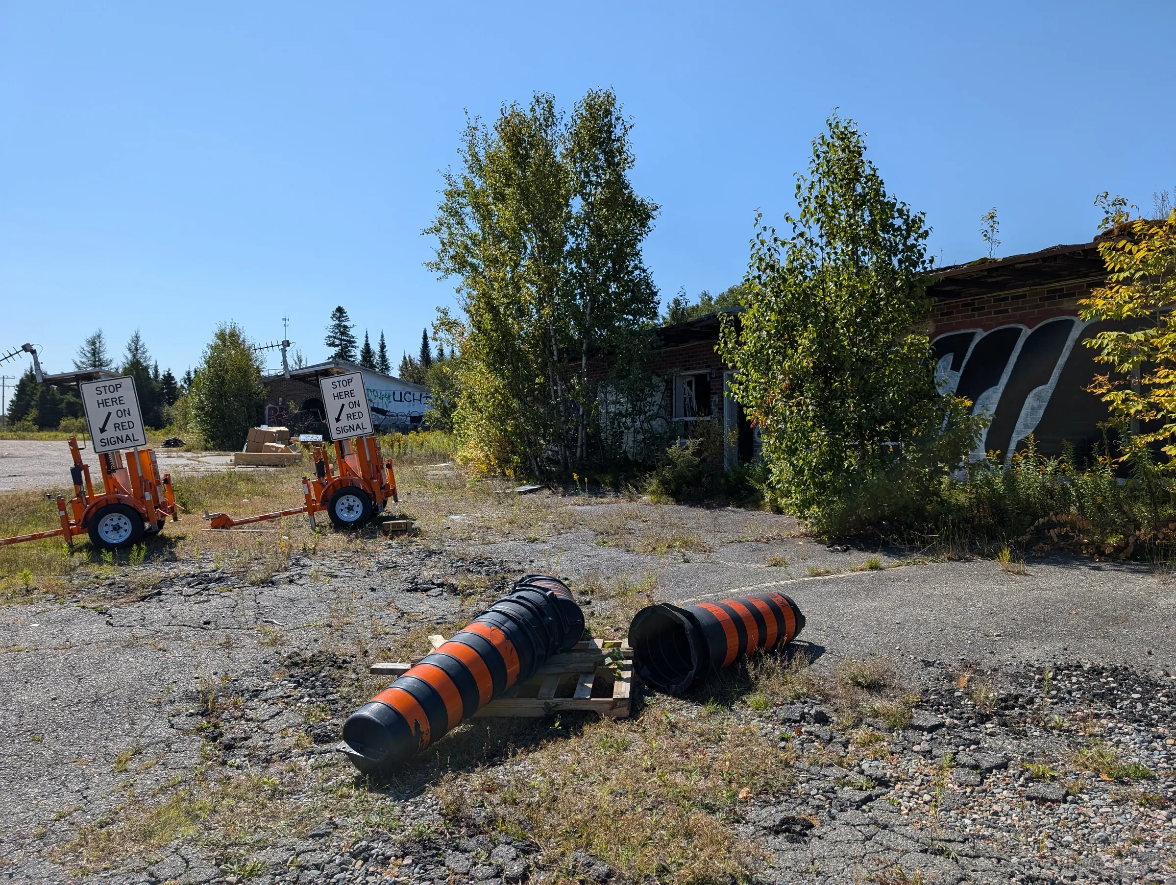 Two portable traffic signal trailers wait at the edge of the lot while toppled orange-and-black barrels lie on a pallet in front of the overgrown, graffitied motel wing.