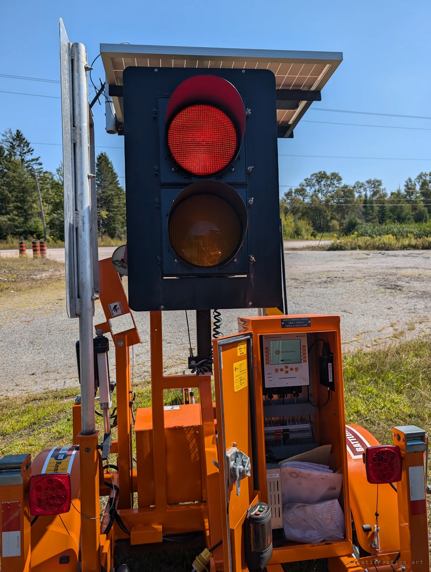 Close up of a portable traffic light glowing red on its orange trailer, the control cabinet open with manuals stuffed inside. A working machine parked among the ruins.