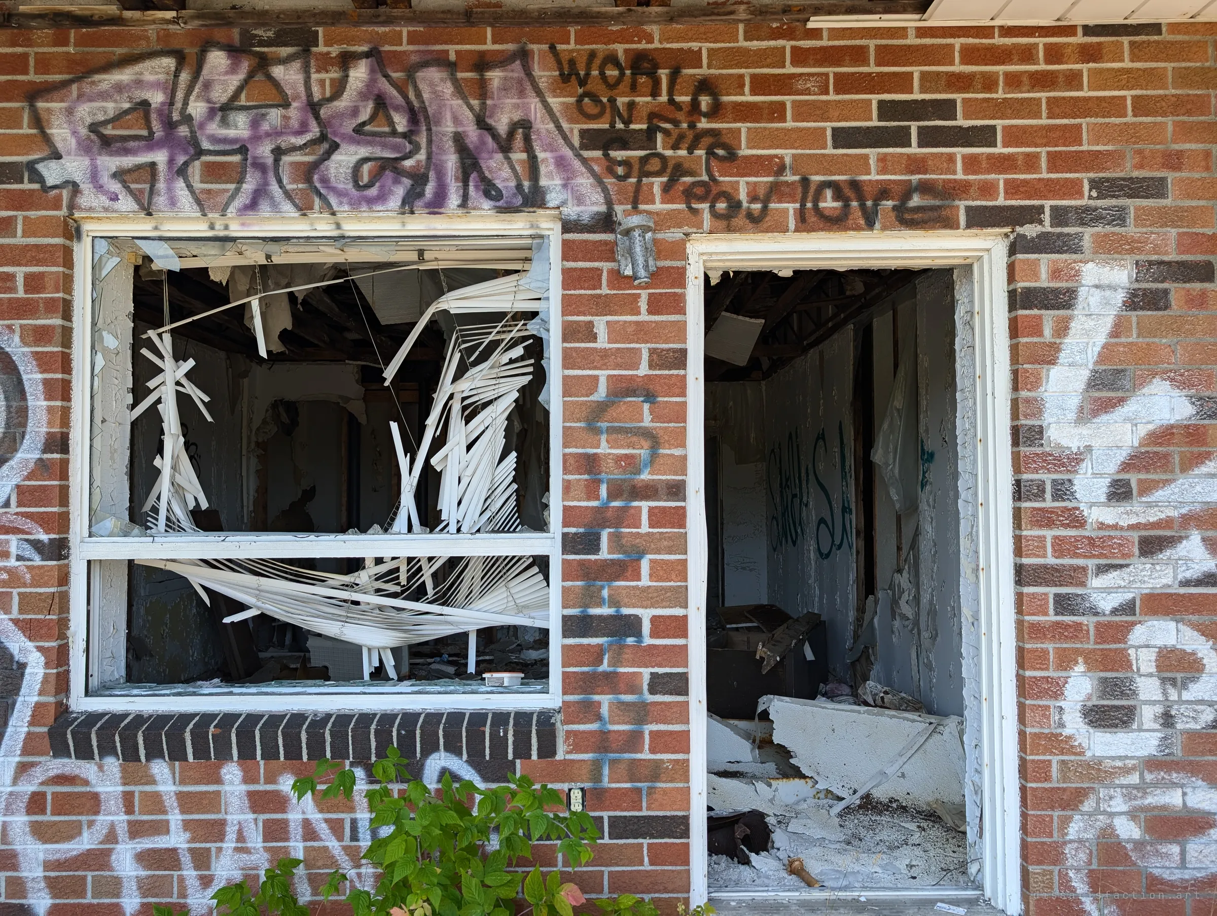A graffiti-covered brick wall with a smashed window full of tangled blinds and an open doorway onto a gutted room. The layered tags show how long the building has sat open.