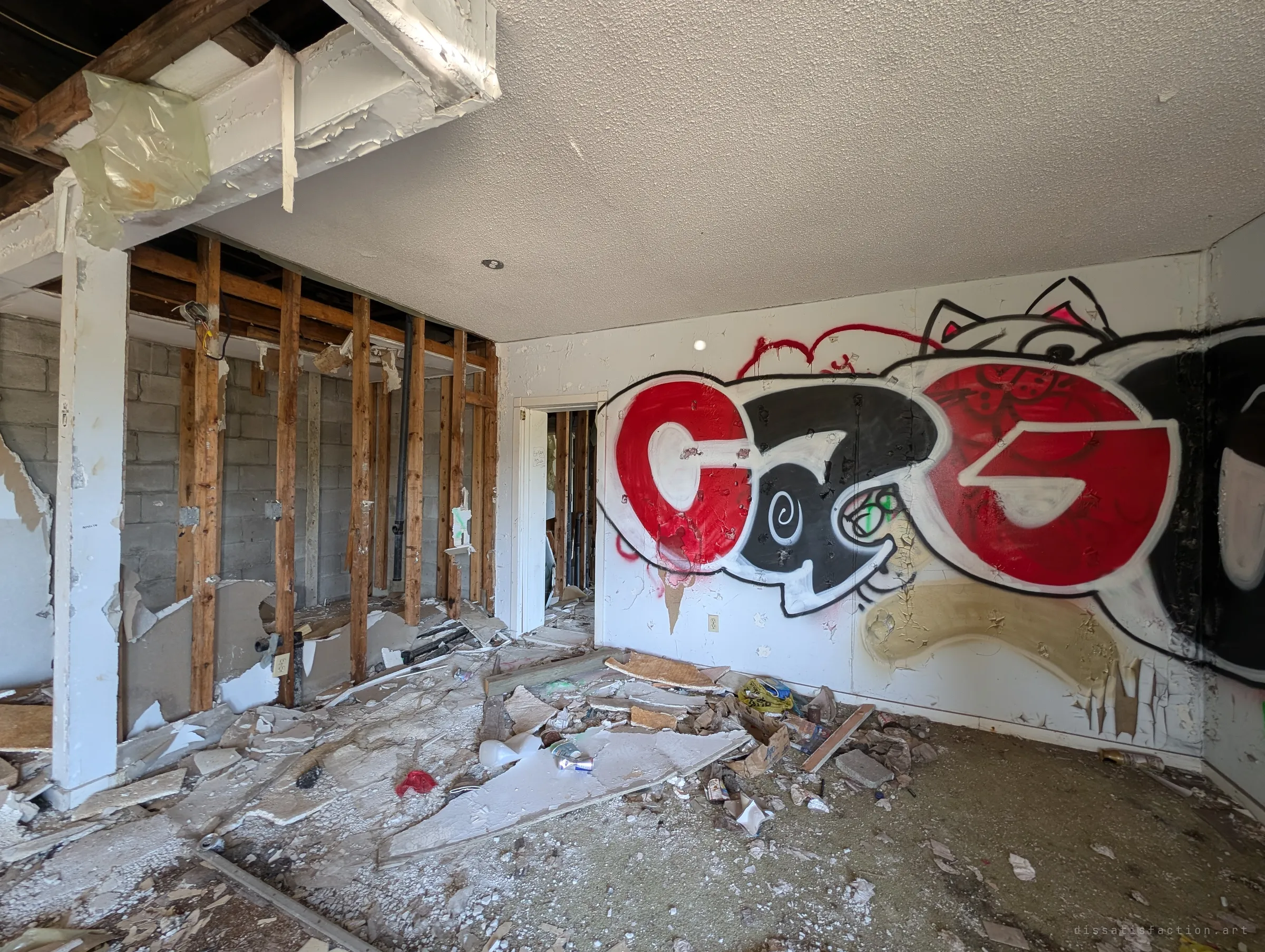 A gutted room with a large red and black graffiti piece across one wall and the partition stripped back to bare studs and block. Drywall and trash cover the floor.