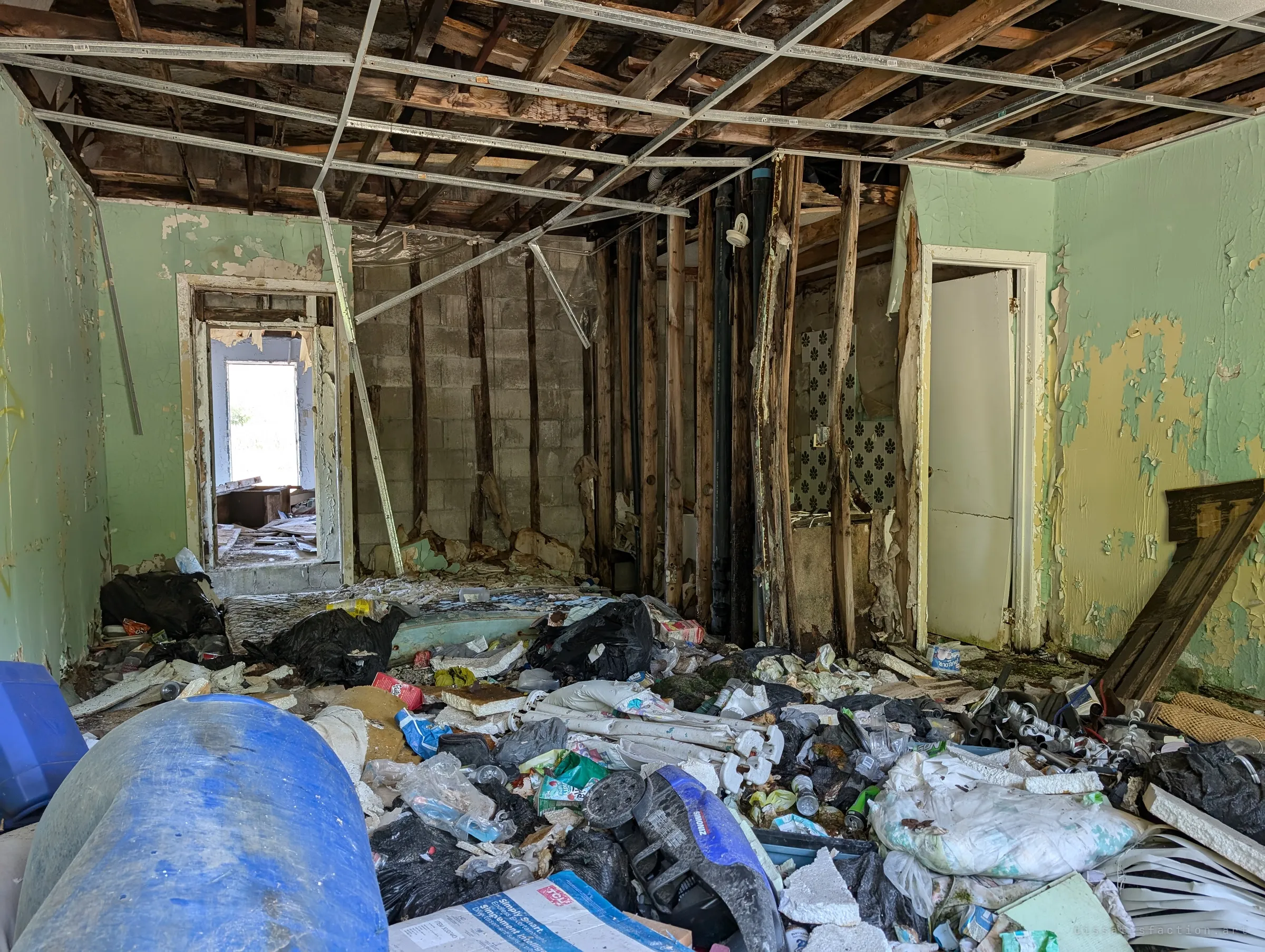 A mint-green room buried in trash bags, bottles and debris under an exposed ceiling grid, with a doorway through to daylight. Rot has eaten the stud wall between rooms.