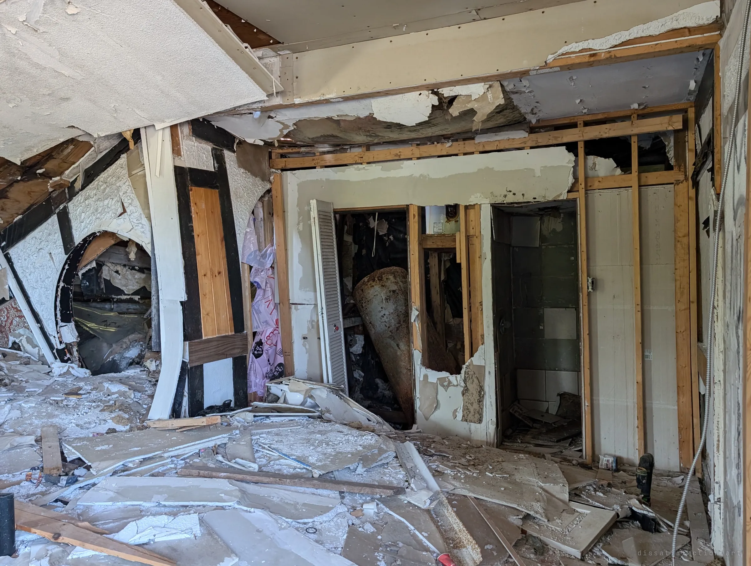 A collapsing interior with an arched opening, half-timbered trim and a toppled water tank wedged in a closet. The ceiling sags down over drifts of broken drywall.