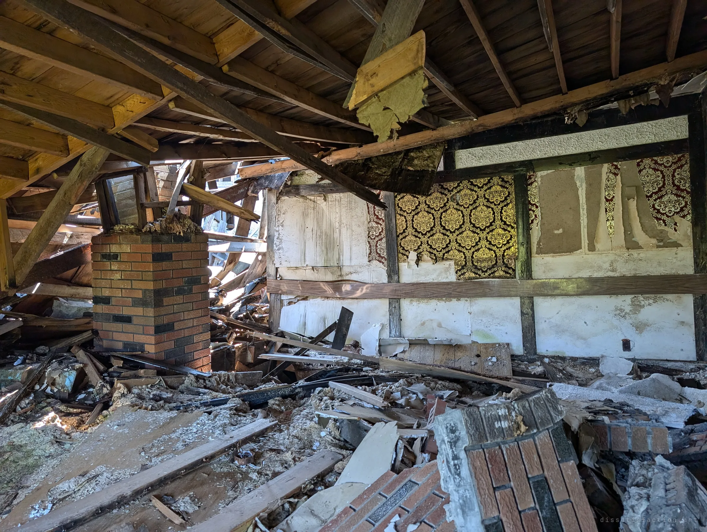 The roof has caved in over a brick chimney stack, leaving joists hanging above walls of ornate gold damask wallpaper. Broken brick and timber fill the floor.