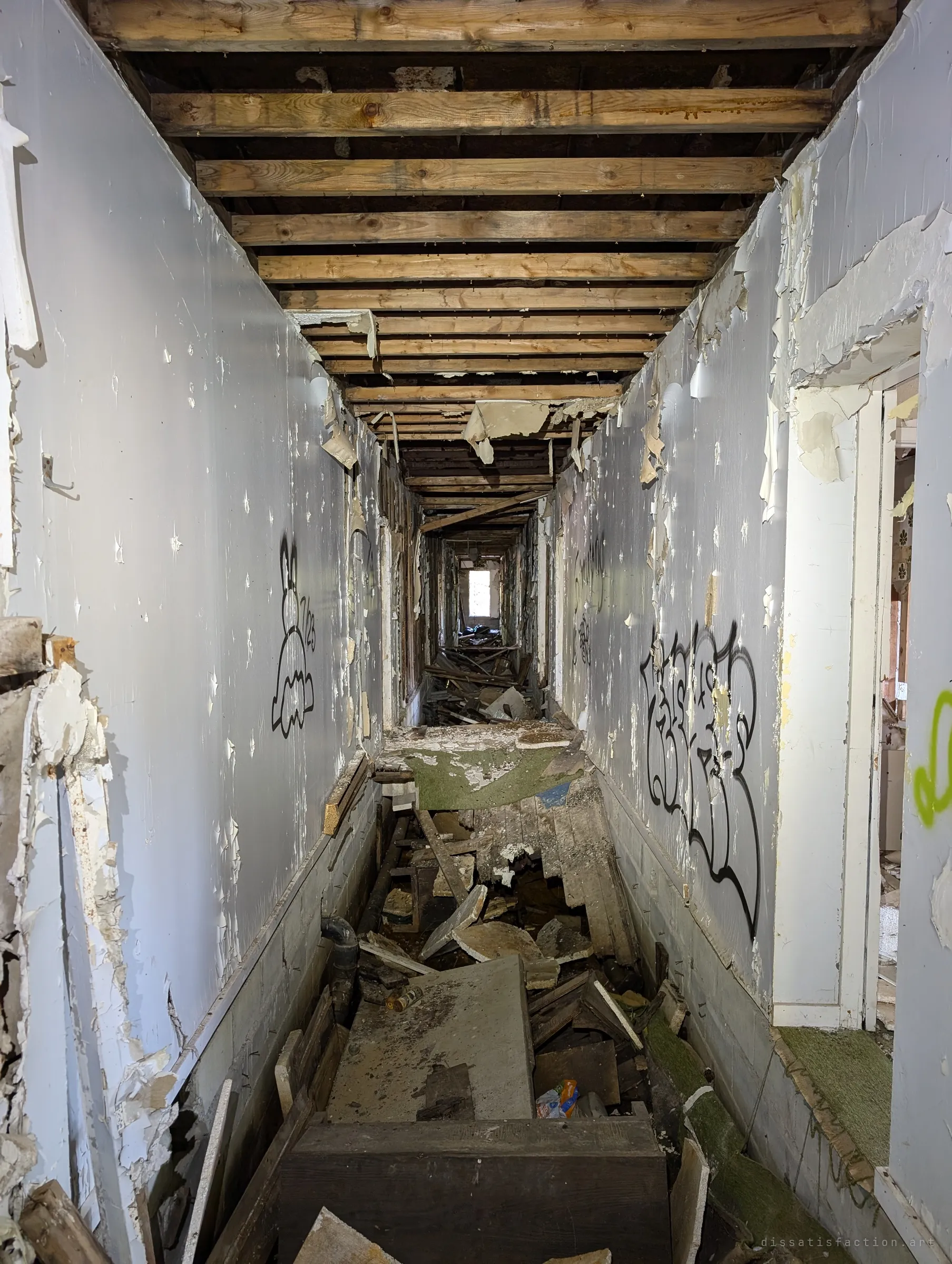 A narrow corridor of peeling white walls and exposed joists runs toward a distant window, its floor broken open into rubble. Graffiti characters line both sides.