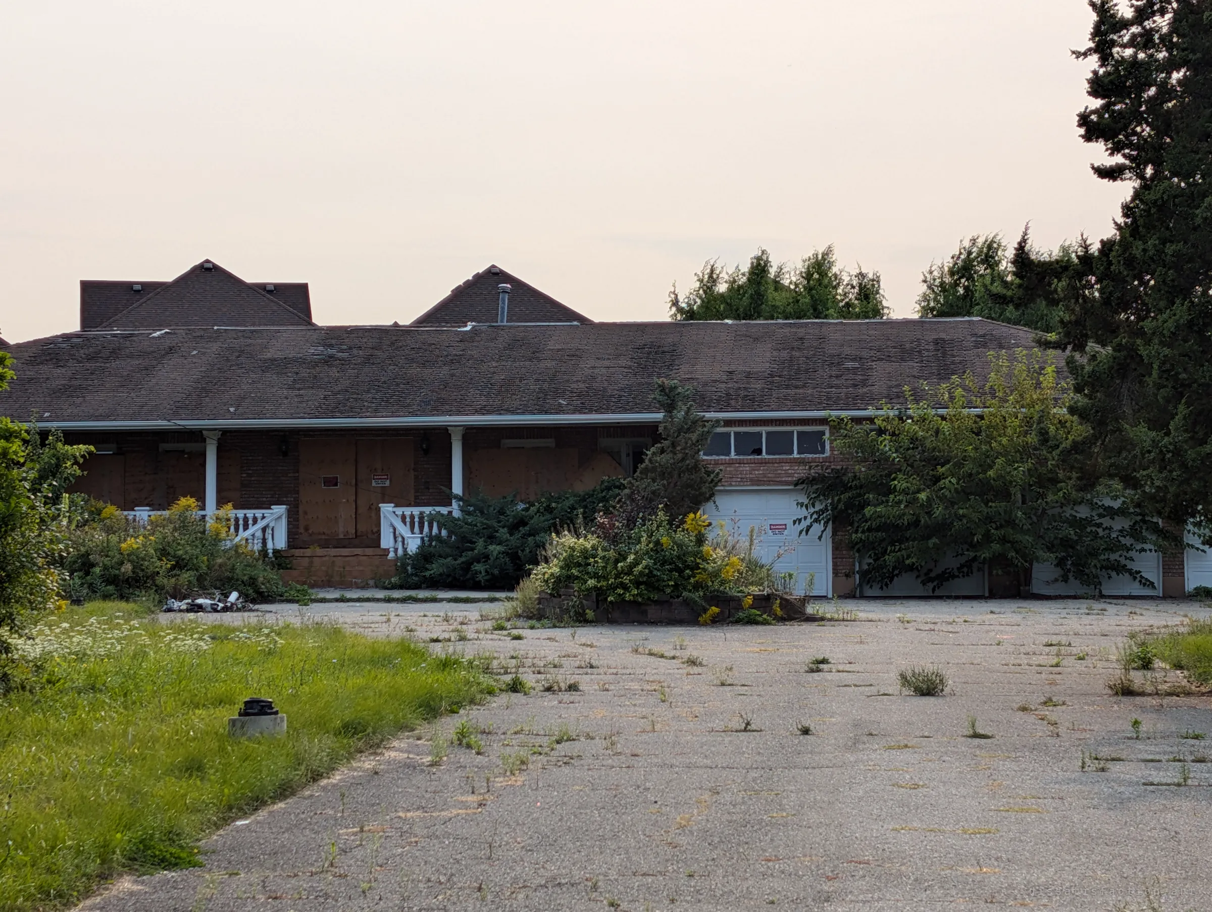 A long brick house with boarded-up doors and windows sits behind an overgrown, weed-cracked driveway under a hazy sky. The warning signs on the plywood set the tone before going inside.