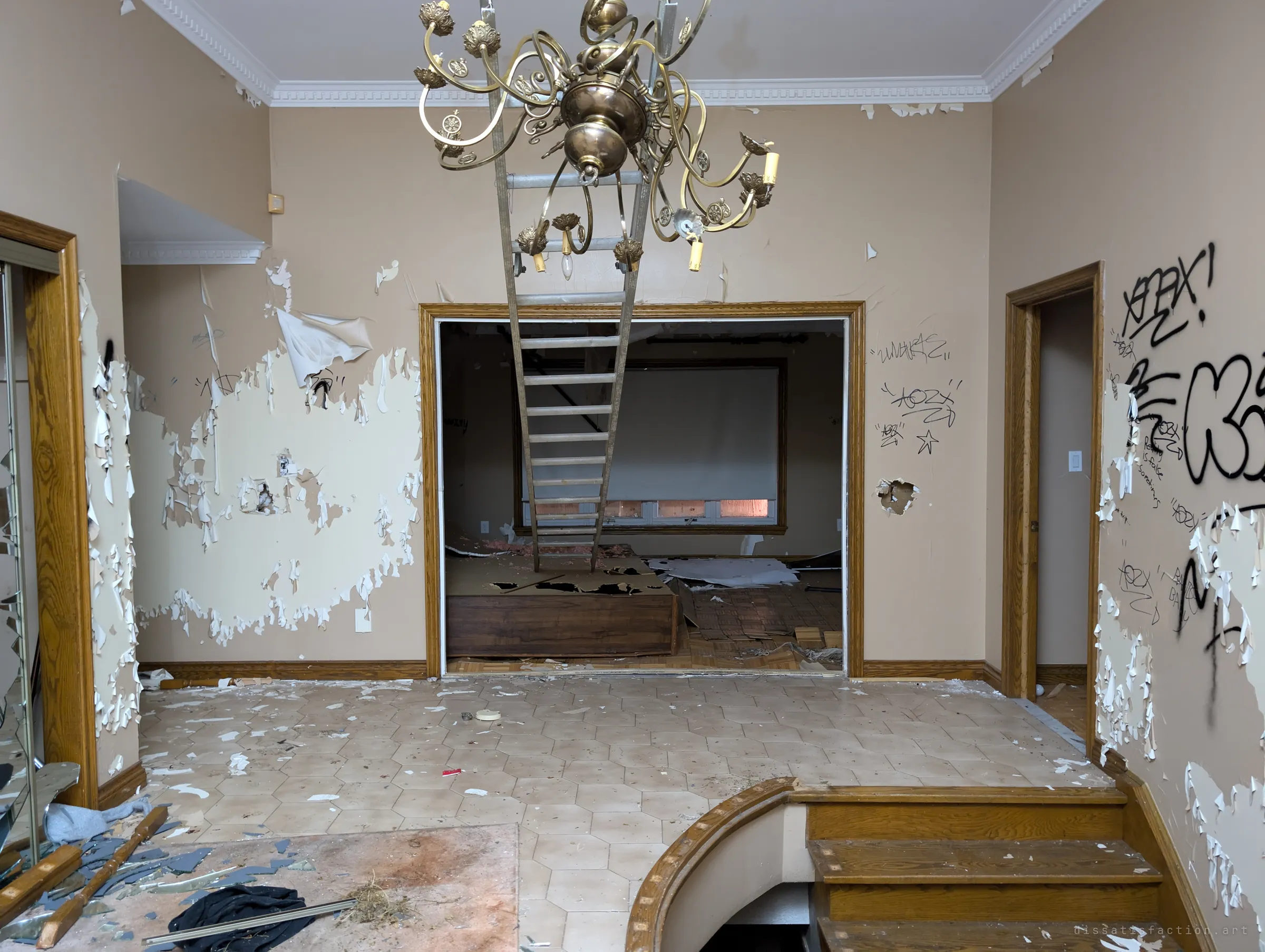 An upstairs landing with peeling paint and graffiti, a brass chandelier hanging in front of an aluminium ladder that leads down into a gutted room beyond. The ladder and the ruined finish show how thoroughly the house has been picked apart.