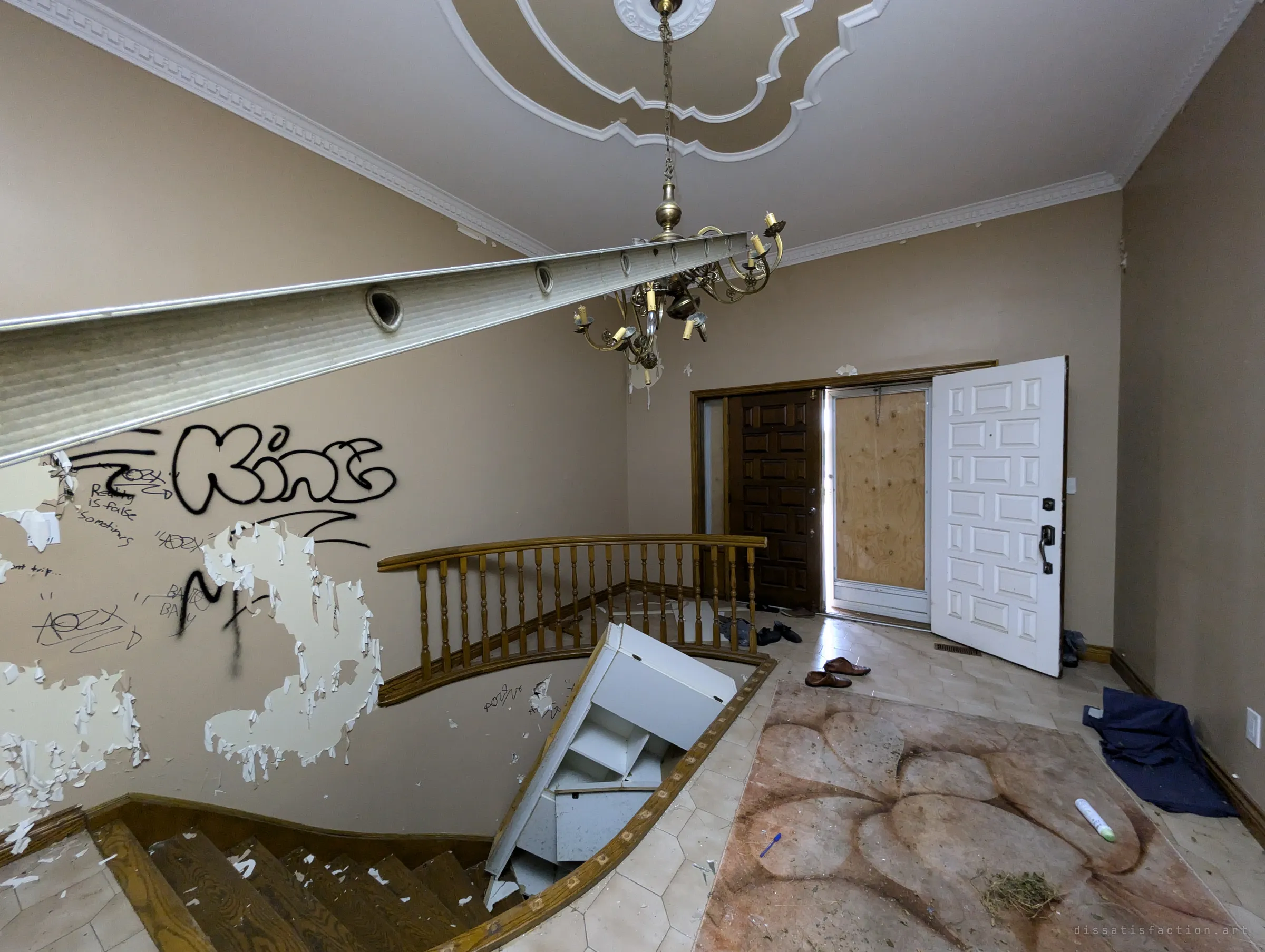 The grand foyer seen from above the curved staircase: an ornate plaster ceiling, a chandelier, a ladder spanning the stairwell and a cabinet tipped into it. The front door stands open onto plywood, with abandoned shoes on the tiles.