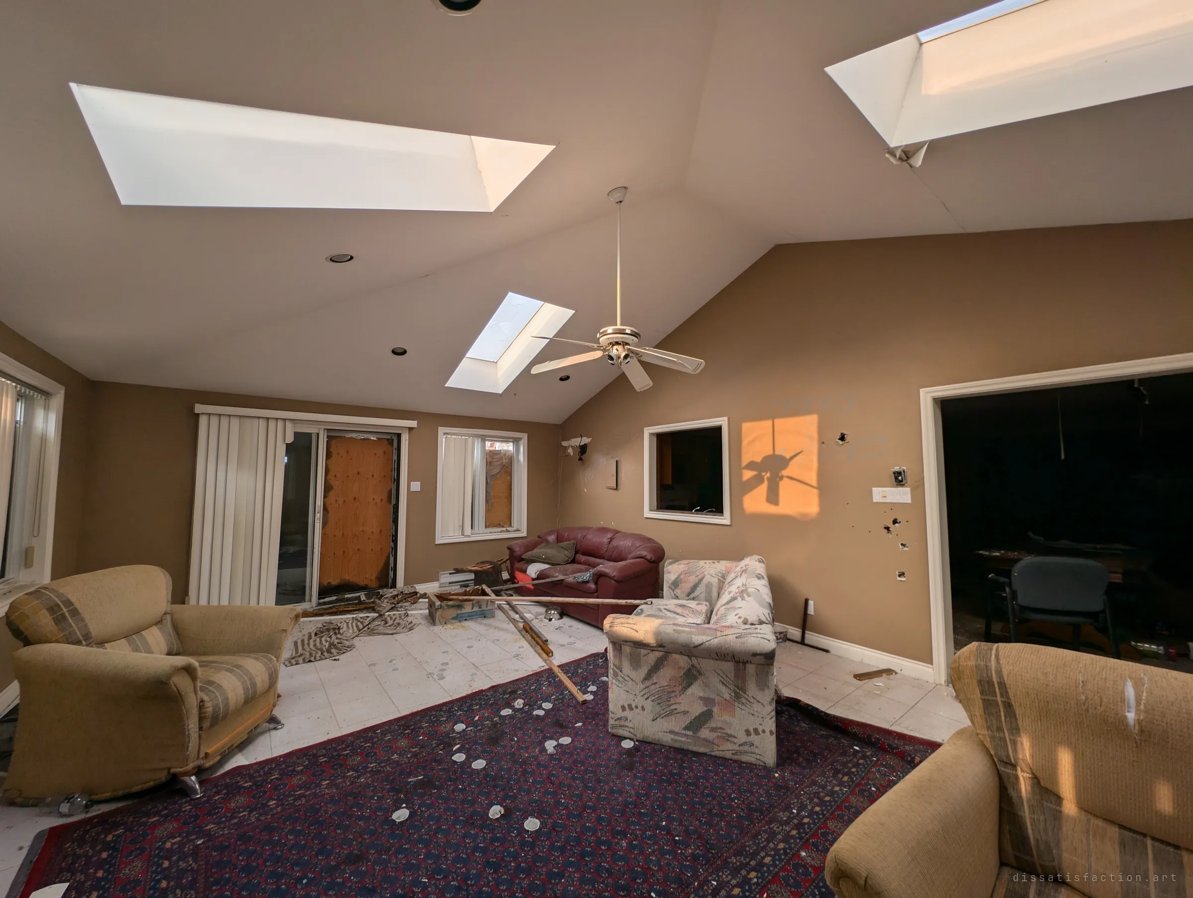 A vaulted sunroom lit by skylights, its armchairs and sofas still arranged around a patterned rug strewn with debris. Late sun throws the ceiling fan's shadow onto the wall beside a boarded patio door.