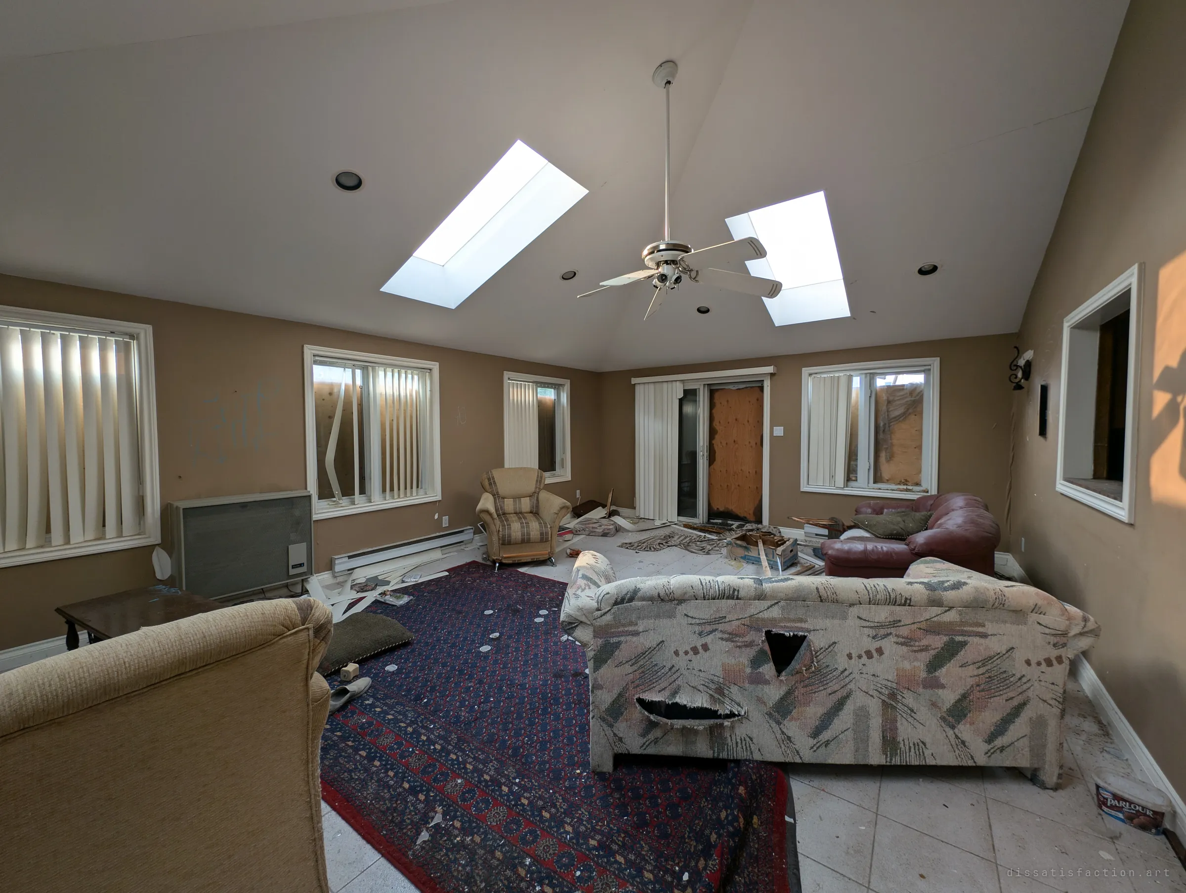 The same skylit sunroom from the other end: a slashed floral sofa, a red leather loveseat and scattered boards across the rug. Every window behind the blinds is sealed with plywood.