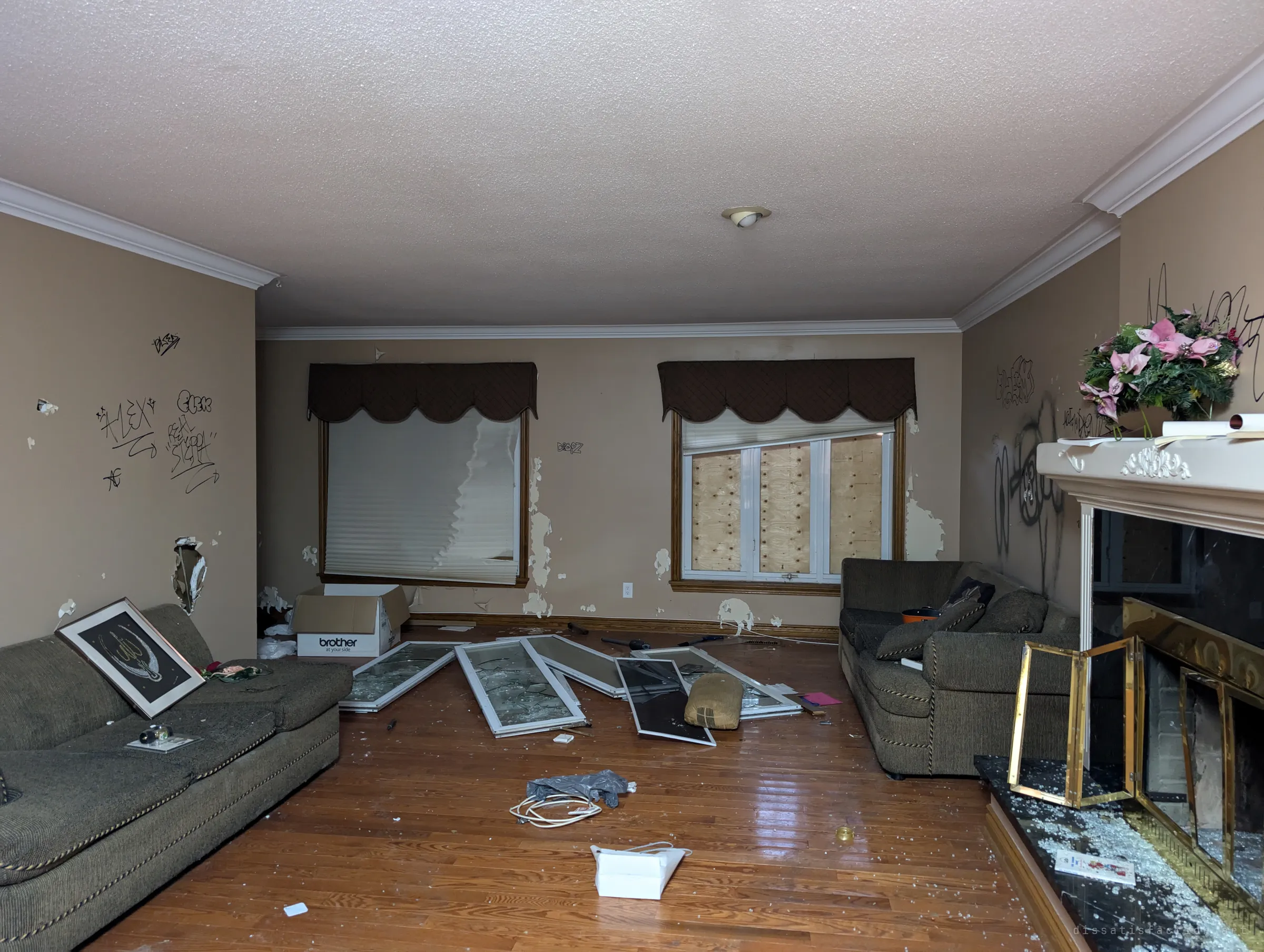 A living room with tagged walls, two sofas facing each other and window frames smashed across the hardwood floor. A dusty flower arrangement still sits on the mantel above a fireplace full of broken glass.