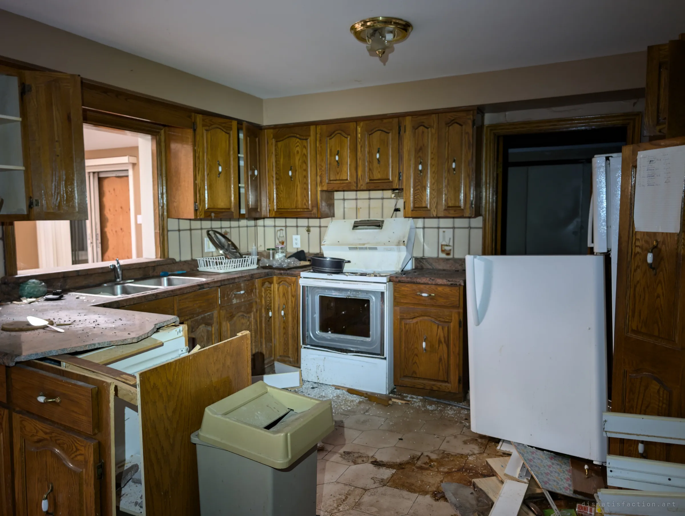 A wood-panelled kitchen left mid-life: a pot on the stove, dishes in the rack and the fridge door torn off and propped against the counter. Broken glass and water stains cover the tiled floor.