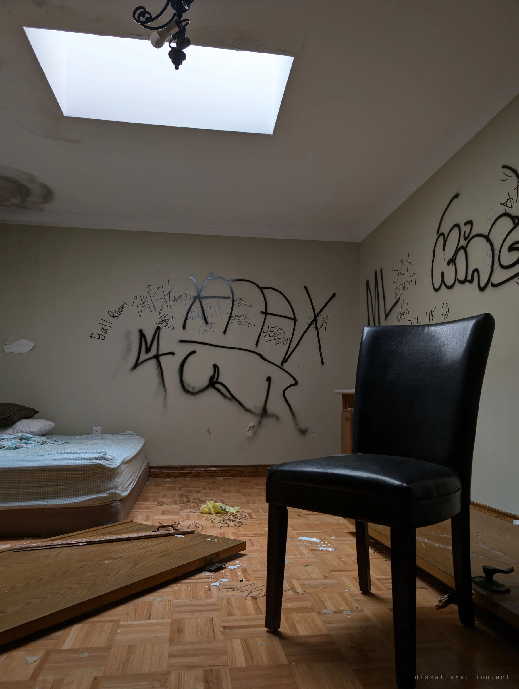 A black leather chair stands alone in a graffiti-covered bedroom under a bright skylight, a bare mattress pushed against the wall. The tags and a fallen door on the parquet show how often the house has been visited.