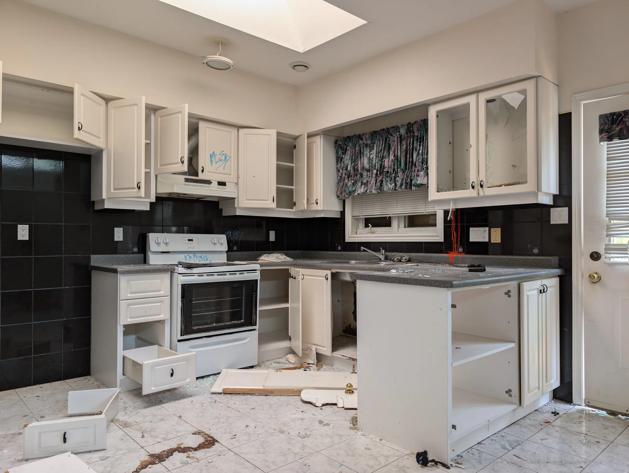 A trashed white kitchen under a skylight, cabinet doors hanging open, drawers and broken glass across the tiled floor and a drip of red paint down the black backsplash. The bright, recent finishes make the vandalism feel sudden rather than slow.