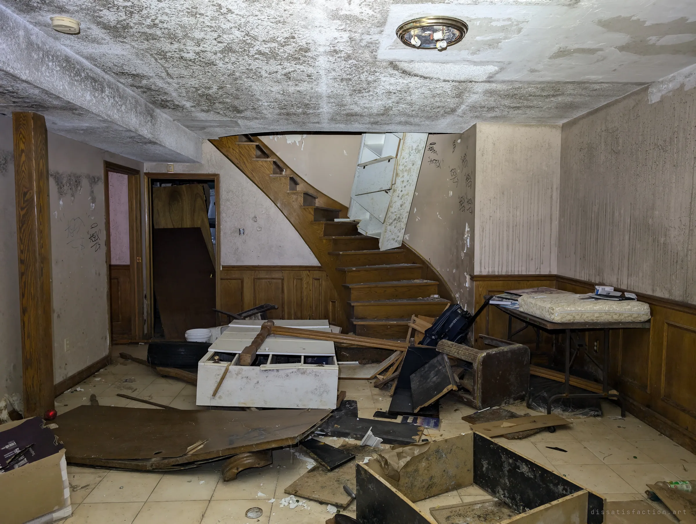 A basement rec room with a curving oak staircase, mould-streaked walls and a ceiling blooming with damp, the floor buried in smashed furniture. Scribbled tags climb the wall beside the stairs.