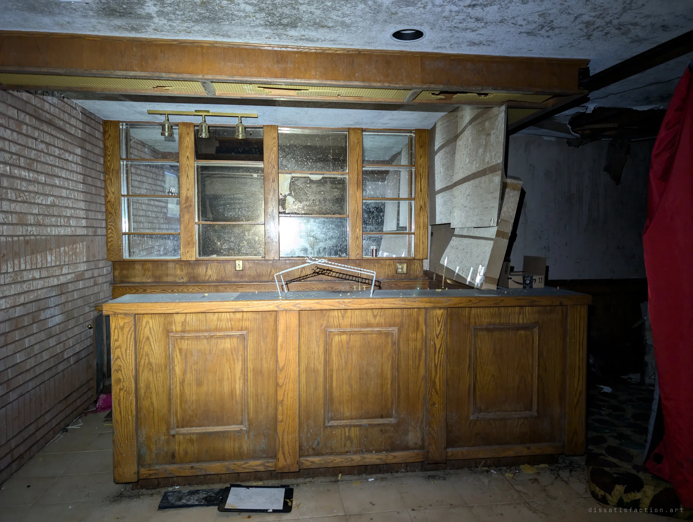 An oak basement bar with mirrored back shelves and brass track lights, lit by a flashlight against the dark. Ceiling panels sag down behind it and a red sleeping bag hangs at the edge of frame.
