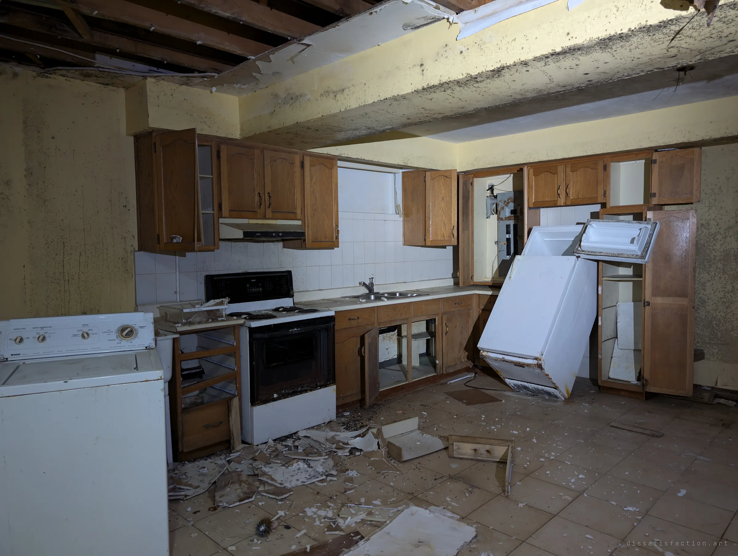 A second, older kitchen in the basement: oak cabinets, a washer, and a fridge tipped over and leaning against the counter under a ceiling torn open to the joists. Black mould spots every wall.