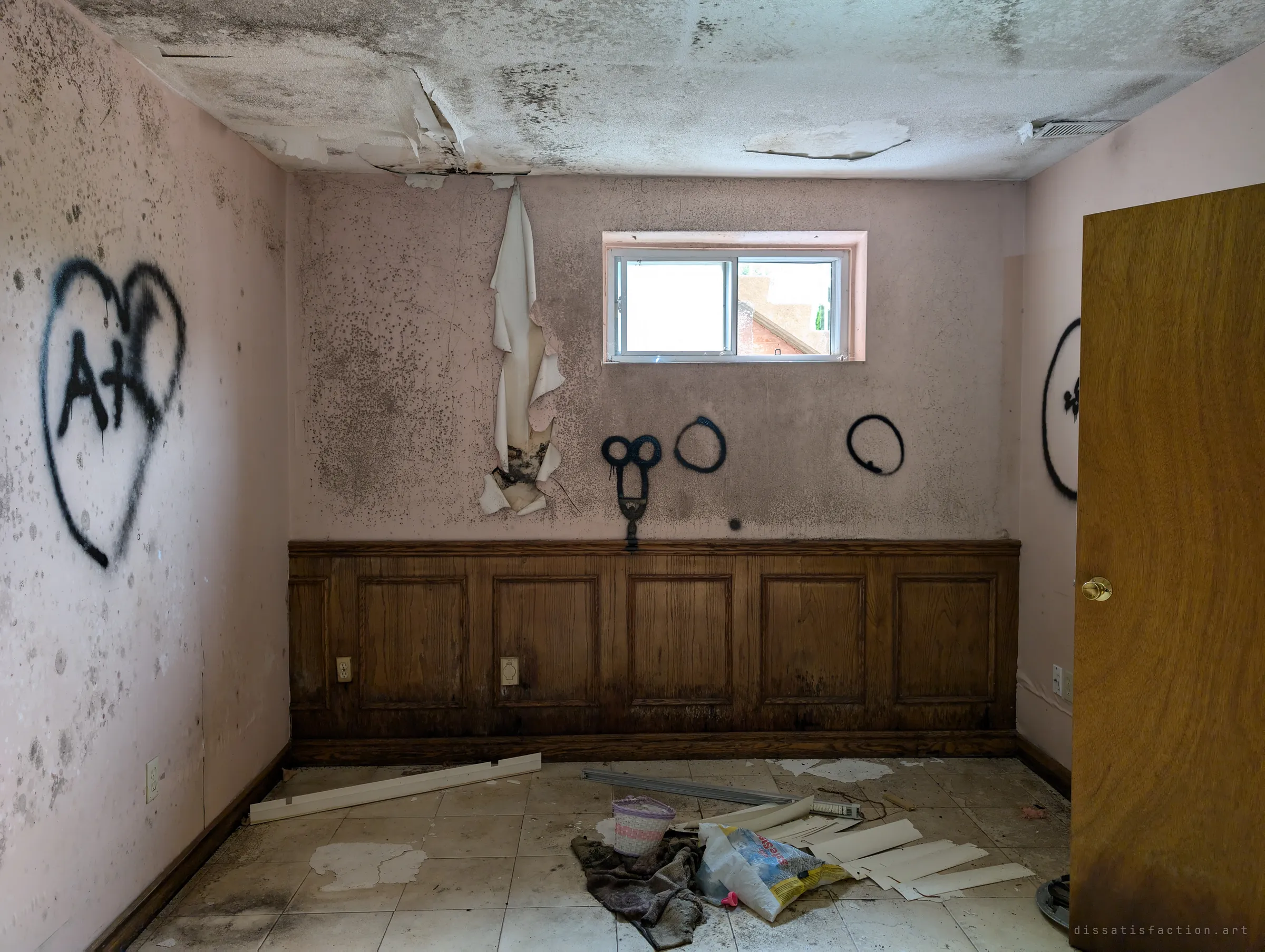 A pink basement bedroom with oak wainscoting, a heart and loops spray-painted on the walls and a strip of wallpaper peeling down from the ceiling. Daylight from a small high window lights the mould.