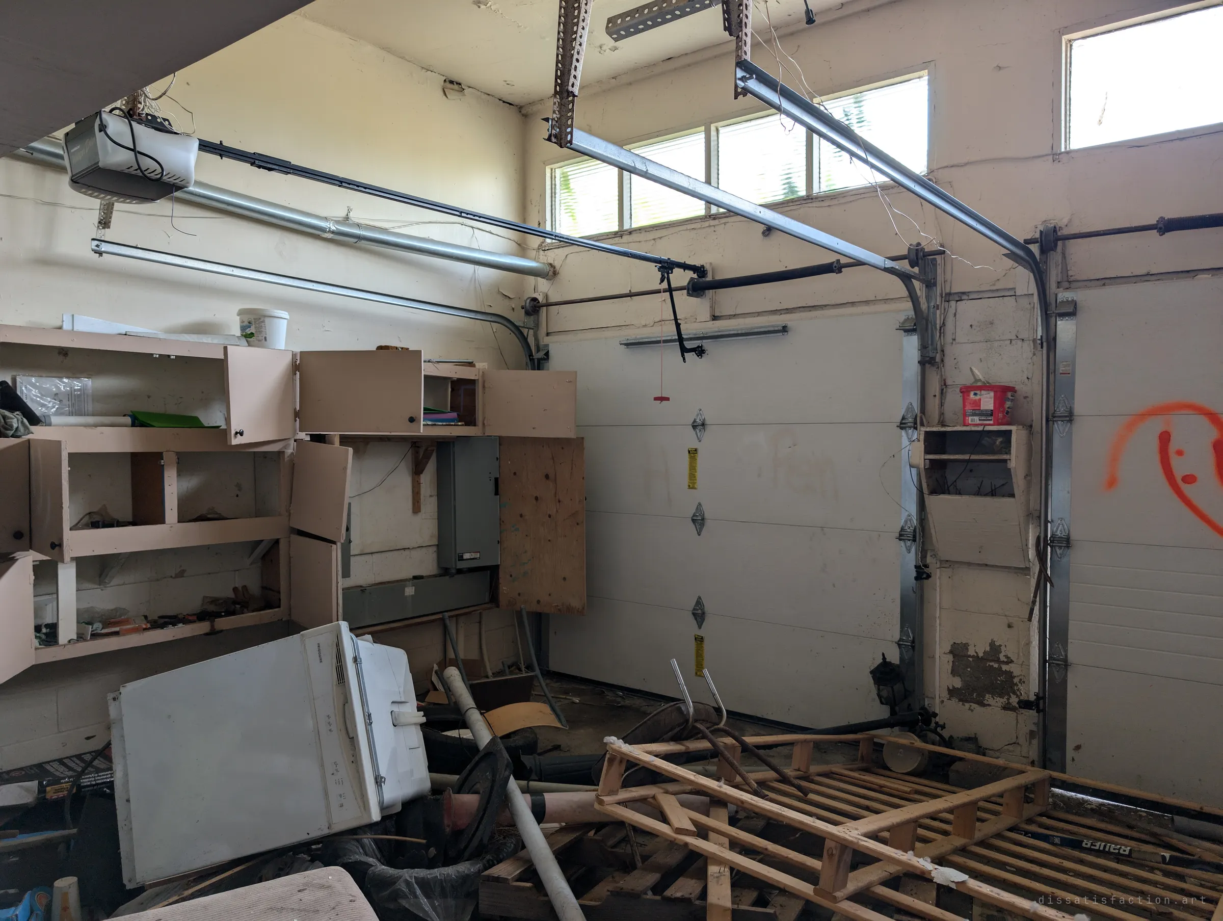 The garage, its door tracks and opener still hanging from the ceiling over a heap of pallets, pipes and a toppled dishwasher. An orange spray-painted face grins from the door on the right.